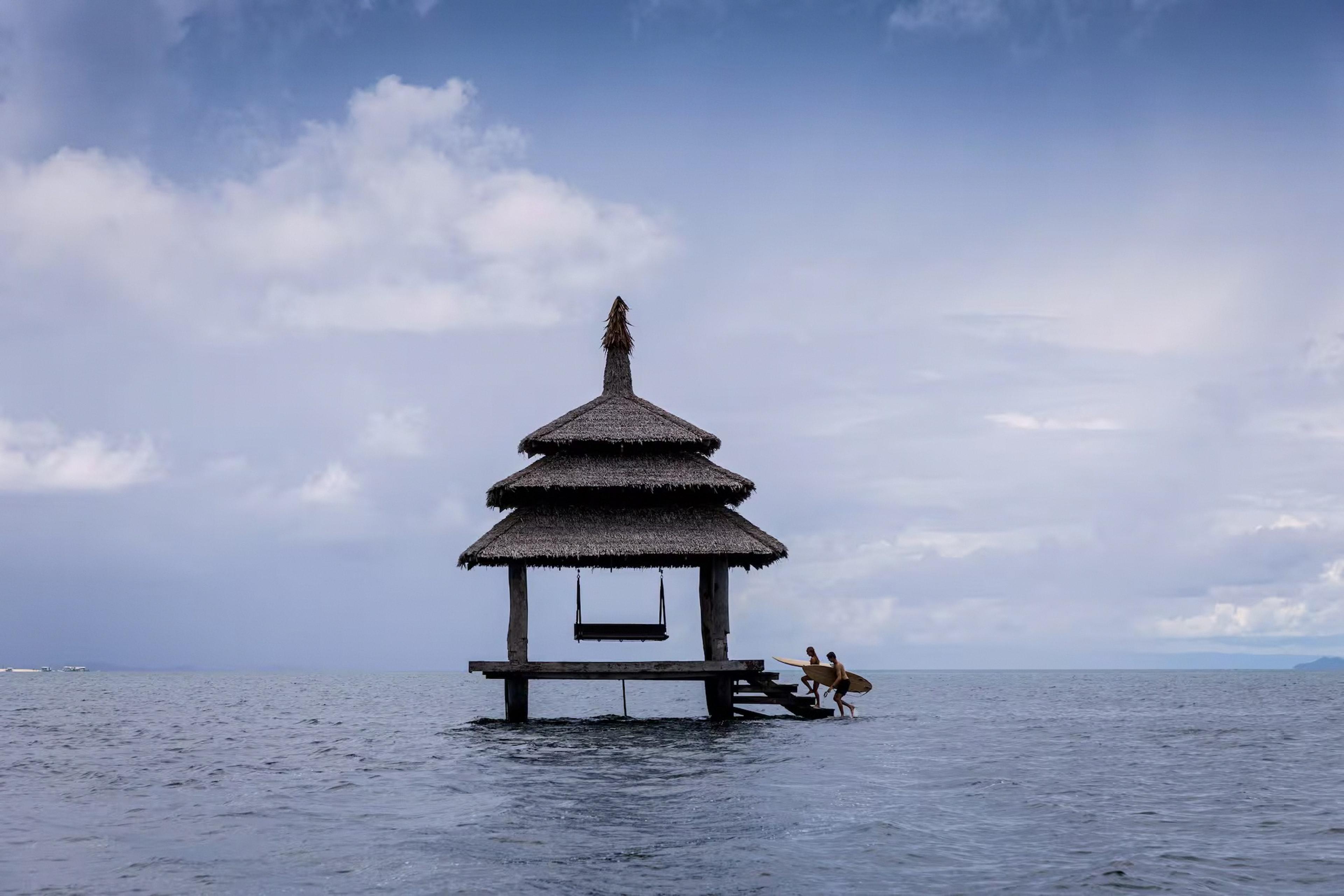 A thatched hut on stilts over calm sea, with two people carrying surfboards walking into the water nearby under a cloudy sky.