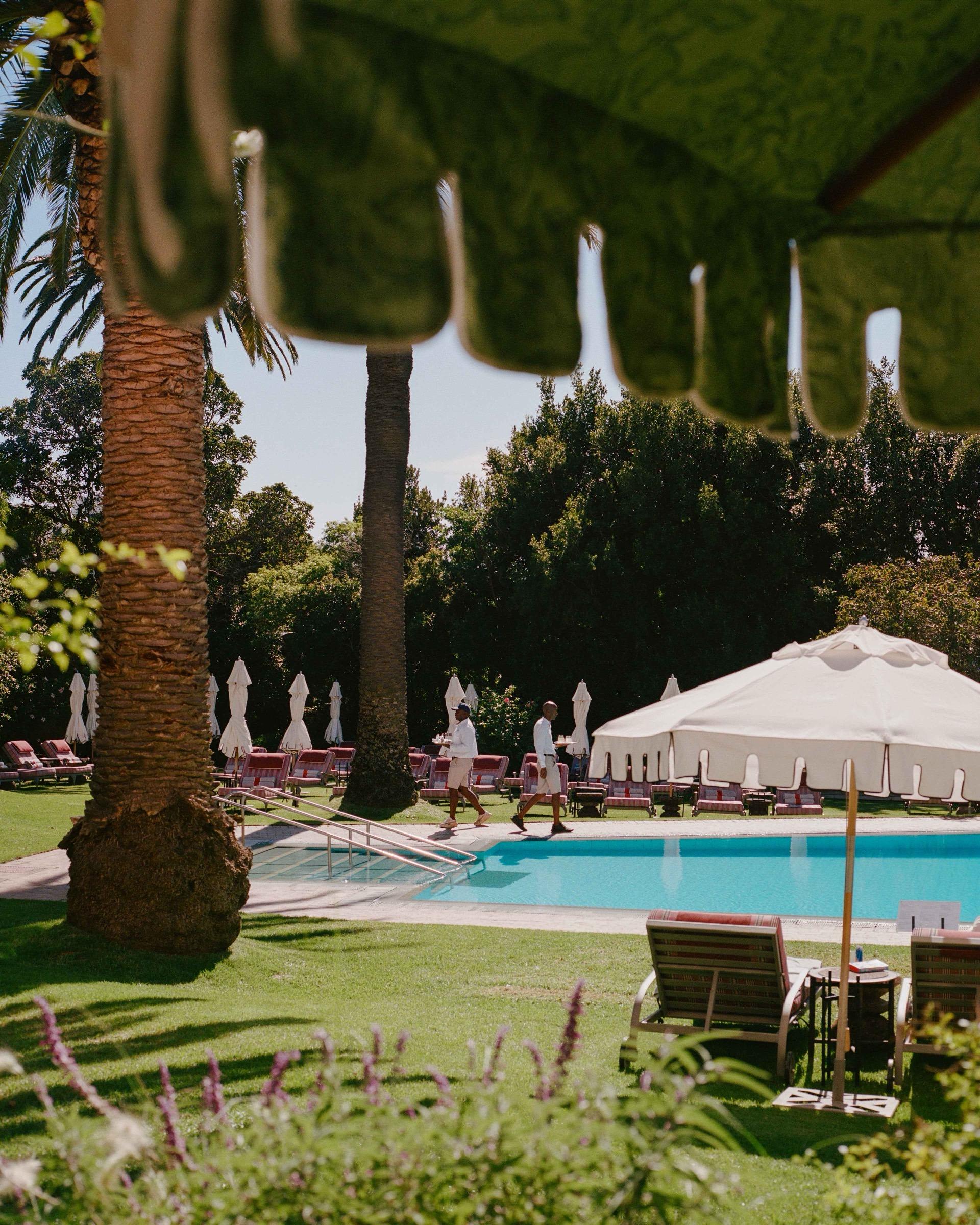 A swimming pool beside a lawn, with white parasols and red loungers, palm trees, and a waiter in white walking past.