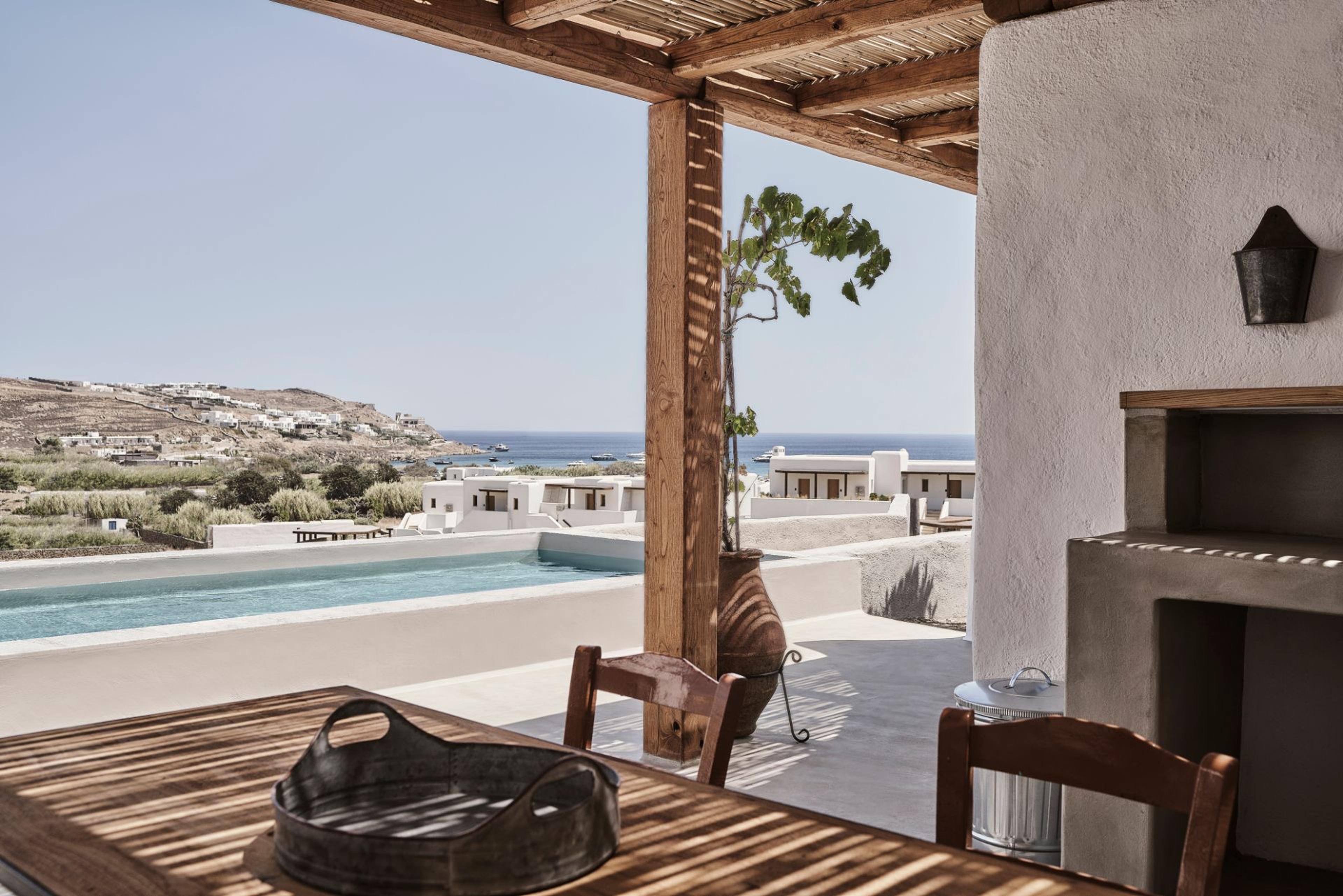 A wooden outdoor dining table with a metal tray sits under a pergola beside a built-in barbecue, looking past a swimming pool toward Mykonos hillside houses and the sea.