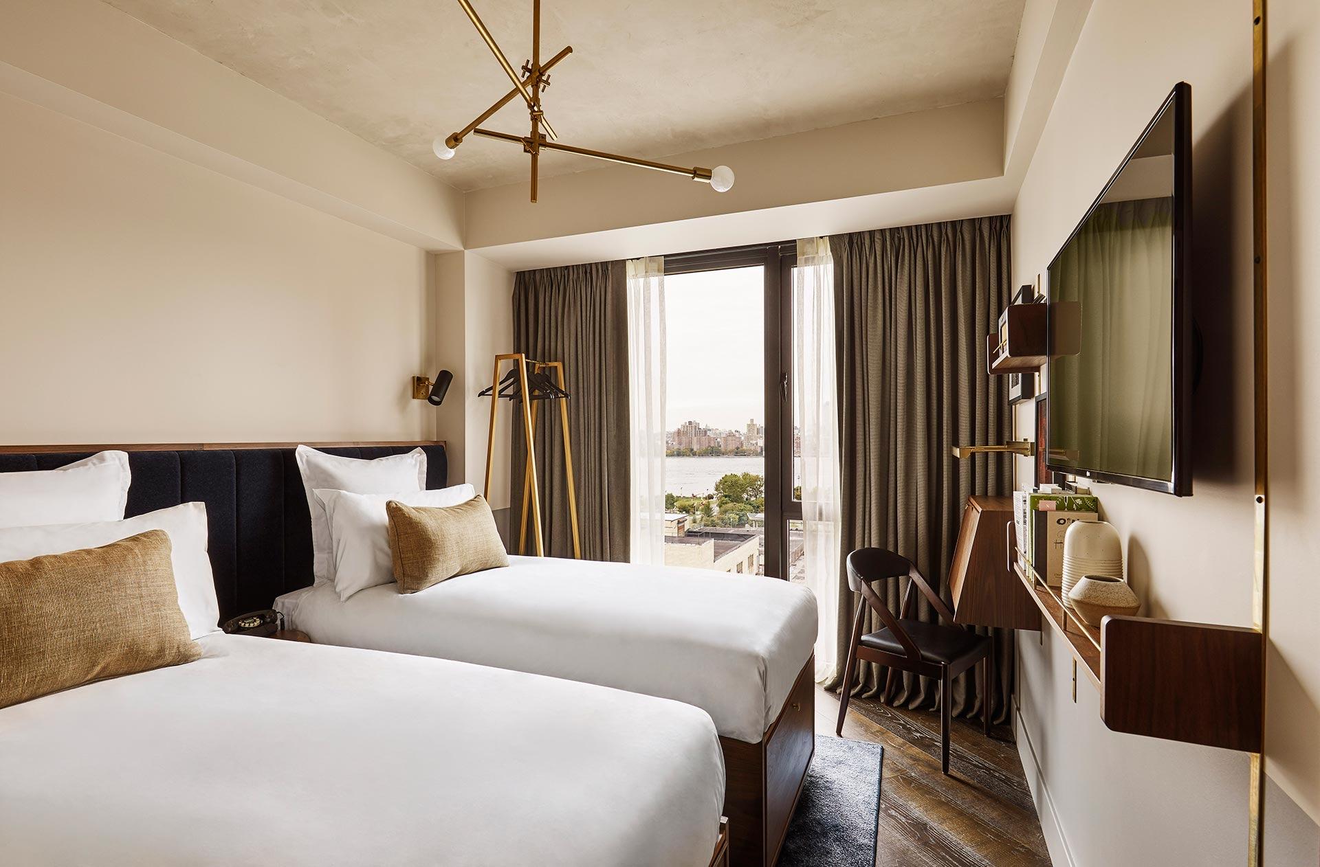 two twin beds under a brass branched light fitting, with a clothing rail and a glass balcony door showing a river view in the distance