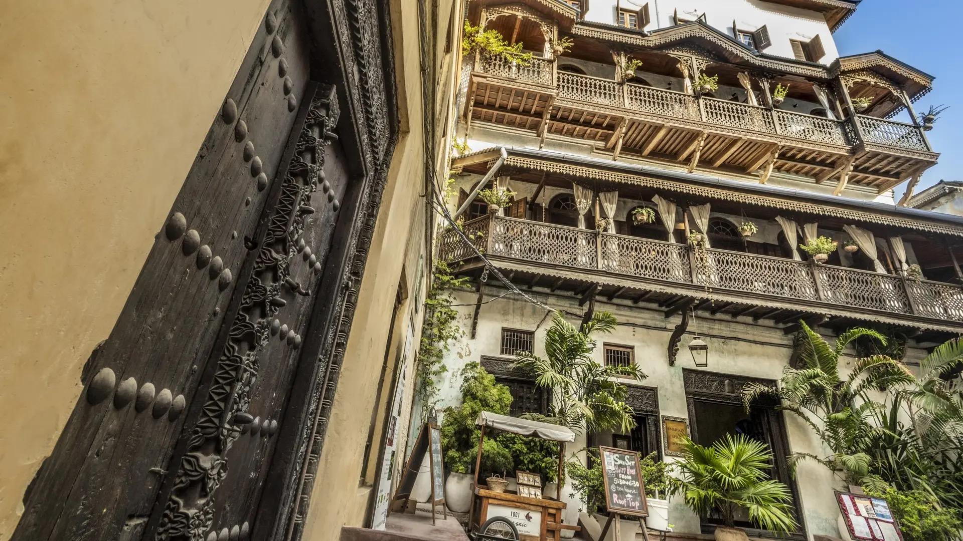 a street view looking up at a tall Zanzibari building with tiered carved wooden balconies, plants and signage on the ground floor