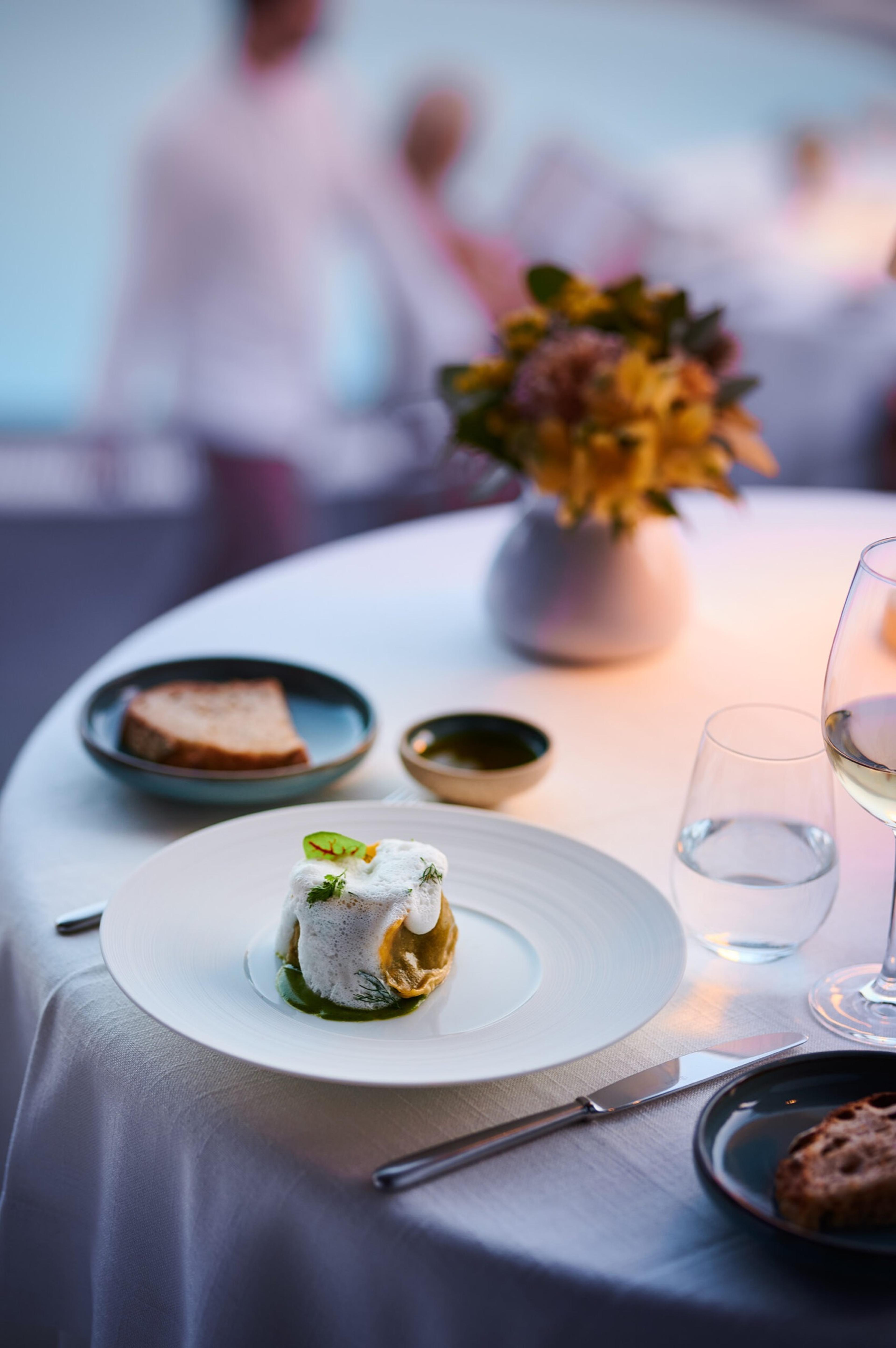 A plated dish with a pastry base and foam topping on a white table, beside bread, a water glass, a wine glass, and a small flower arrangement, with blurred figures in the background.