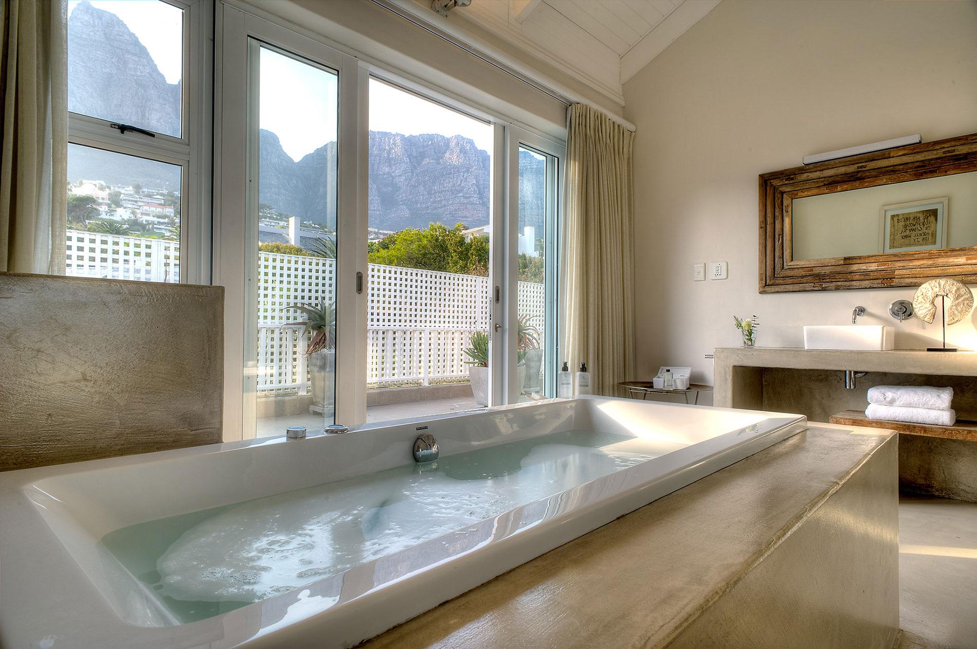A white bath filled with water, set below a window that looks out on a steep rocky mountain and a white balcony rail.