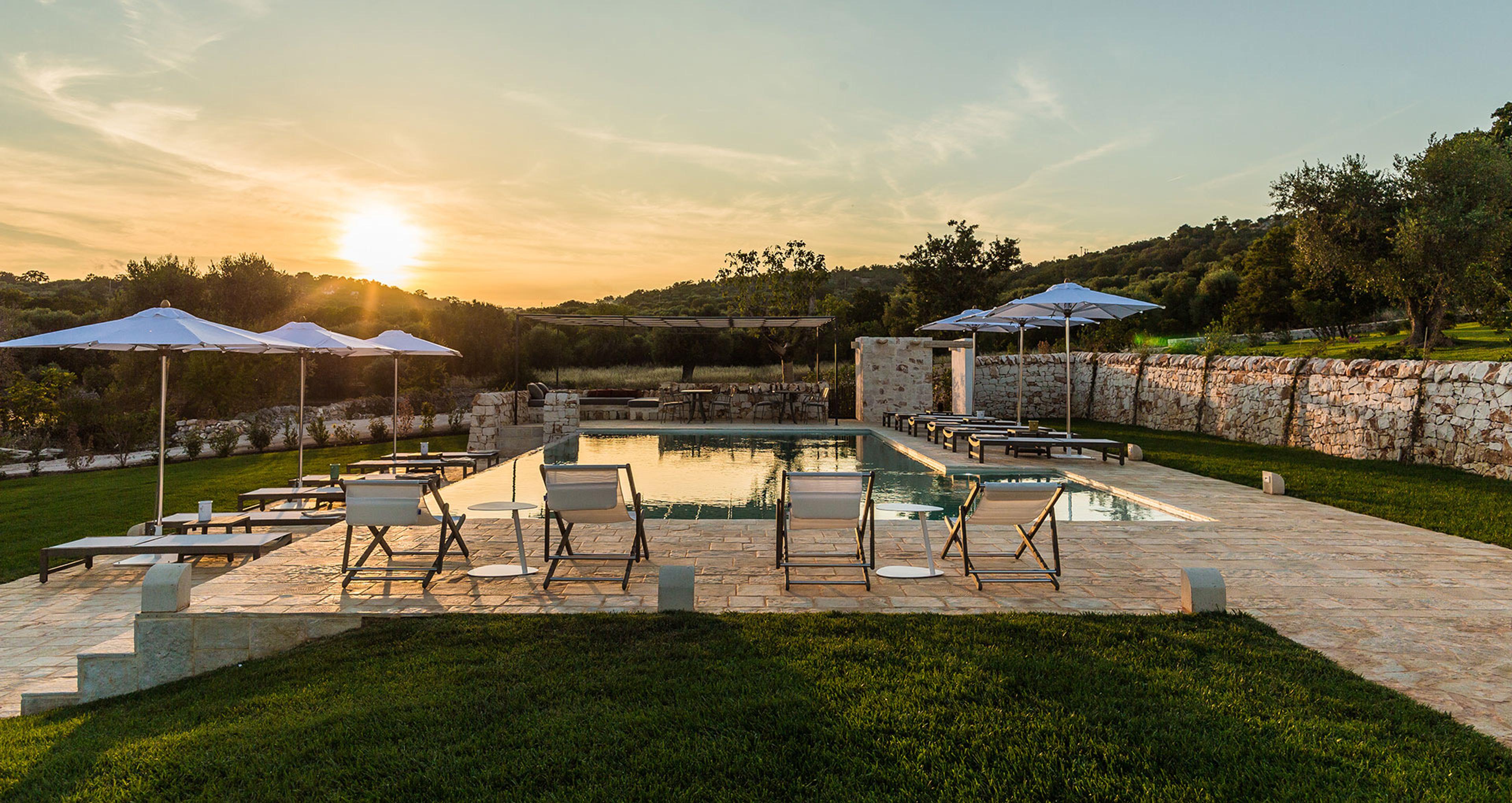 A rectangular swimming pool on a stone terrace lined with sun loungers and blue umbrellas, lit by a low setting sun.