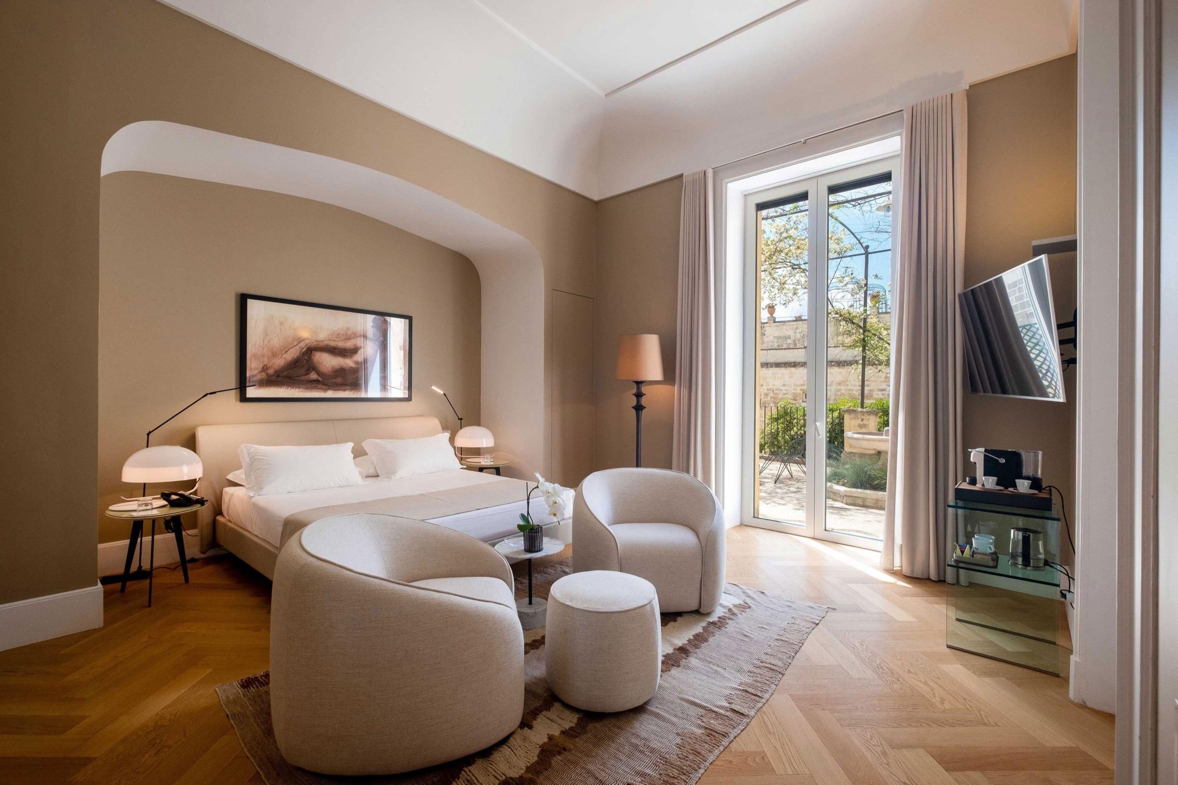 A bedroom with a cream bed set in an arched alcove, two armchairs and a stool, and glass doors open to a small garden terrace.