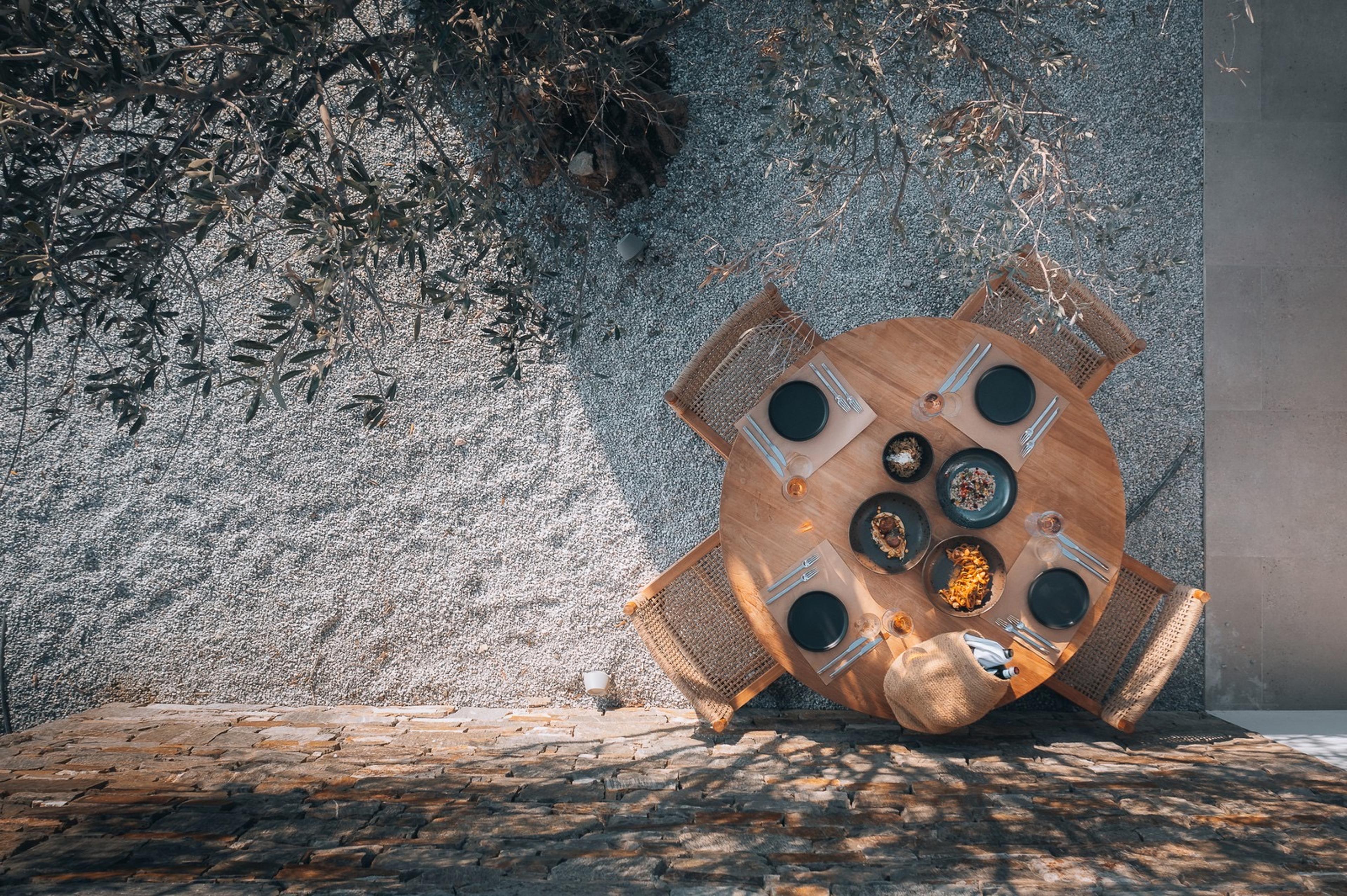 A round wooden table seen from above, set with plates of food beside an olive tree's shadow.