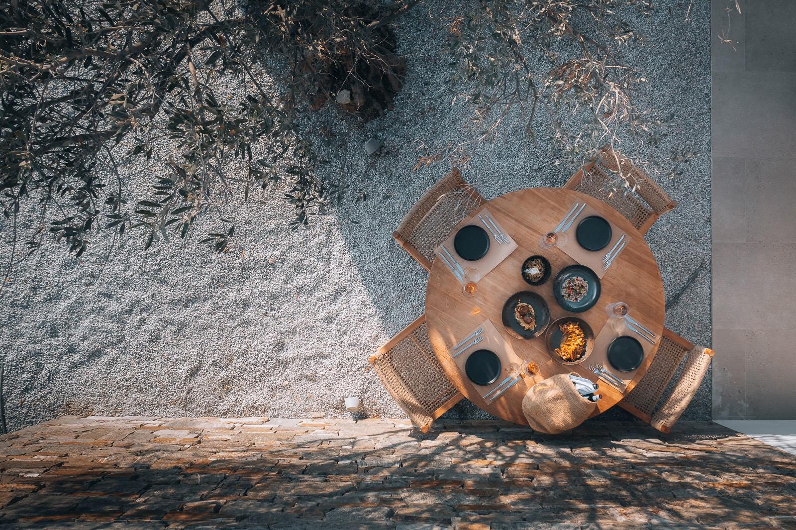 A round wooden table seen from above, set with plates of food beside an olive tree's shadow.