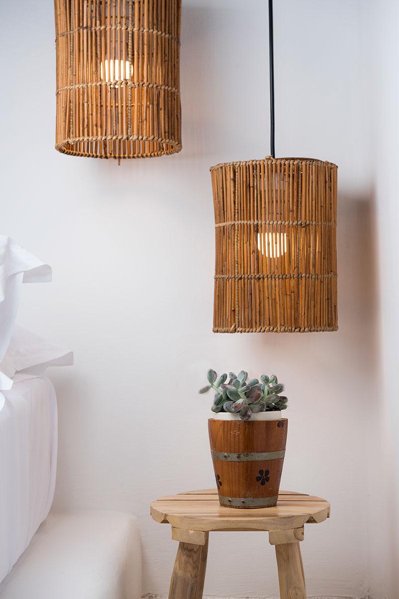 Two woven rattan pendant lamps hang over a bed with white linen, beside a round wooden stool holding a potted succulent.