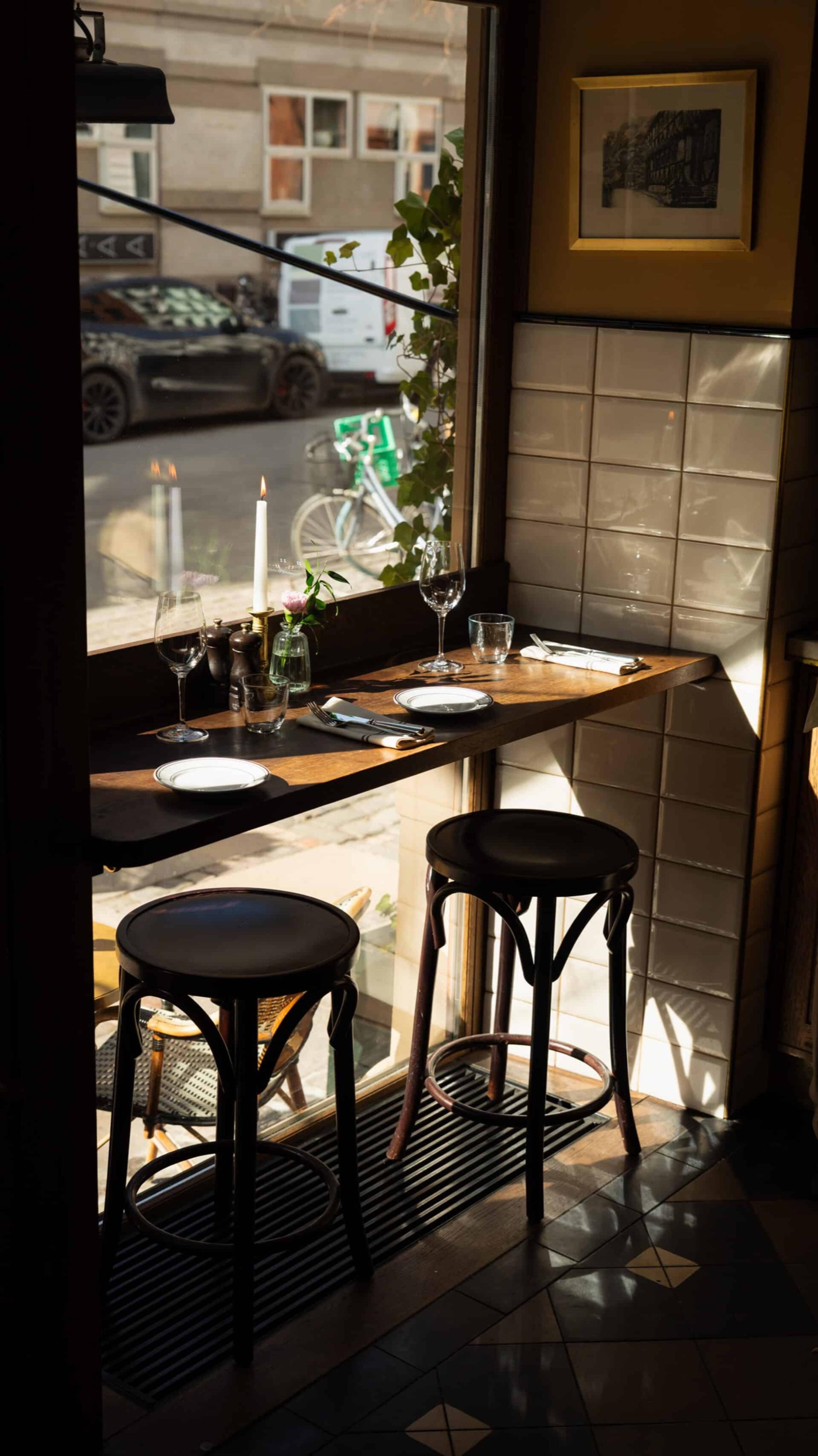 A small window table laid for two with candles and wine glasses, looking out through glazed doors onto a city street with parked cars and a bicycle.