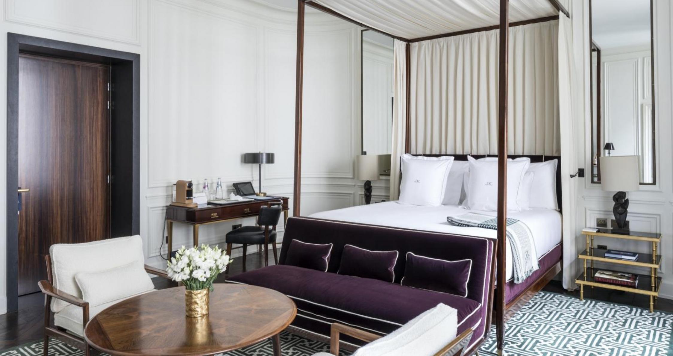 A four-poster bed with a dark wood frame and cream curtains, a deep purple velvet daybed at its foot, and a round table set with flowers.