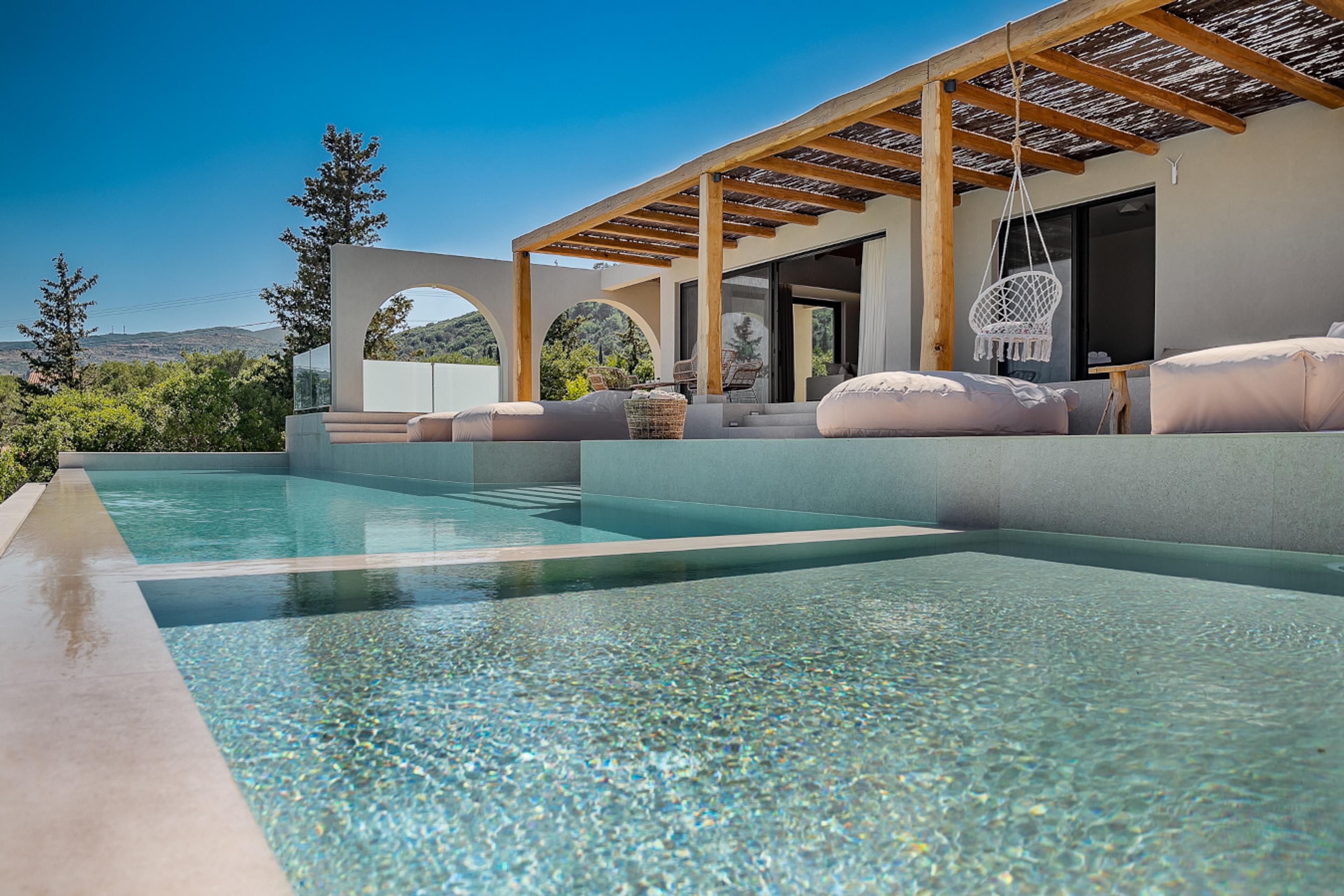 The rippled surface of an infinity pool in the foreground, with the villa's pergola, swing chair and loungers beyond.