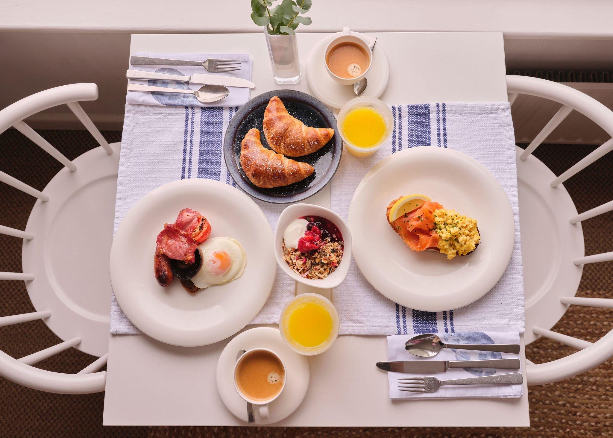An overhead view of a breakfast table laid for two, with a full English, smoked salmon and eggs, granola, croissants, coffee and orange juice.