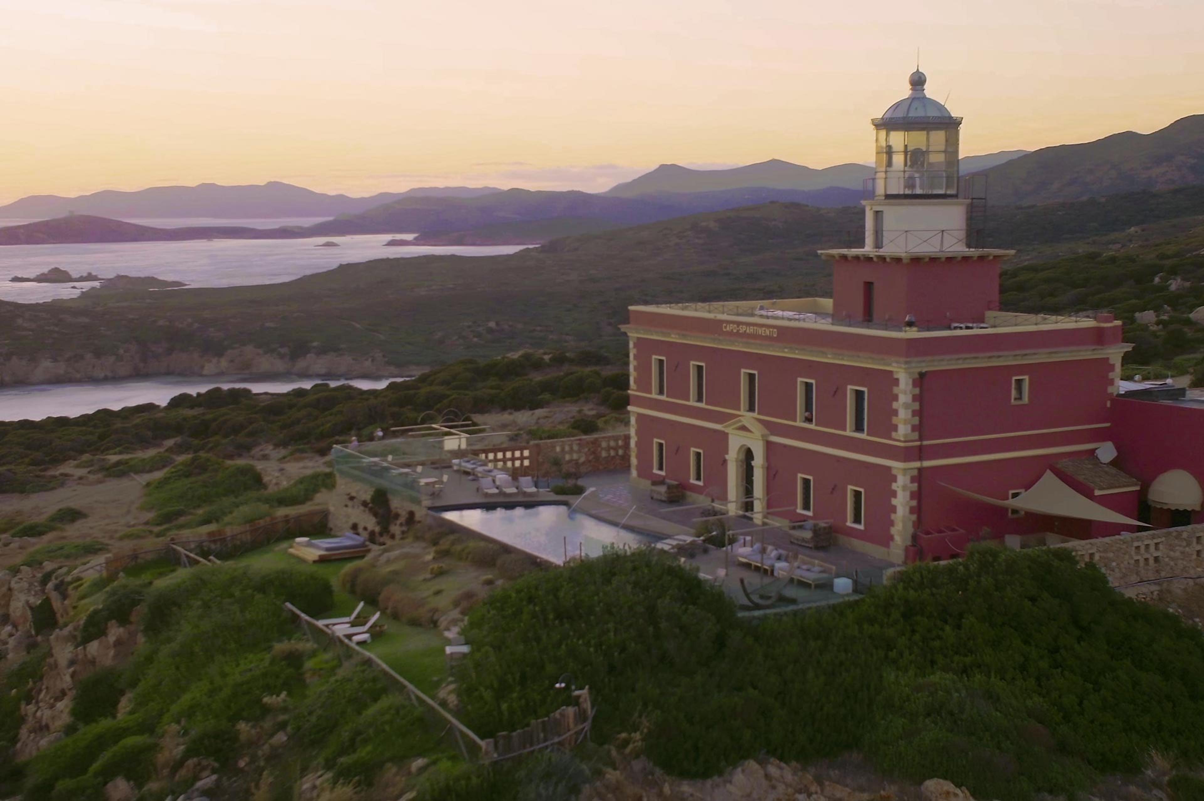 an aerial view at dusk of a red lighthouse keeper's building on a headland, with a pool and terrace below it, the sea and mountains behind