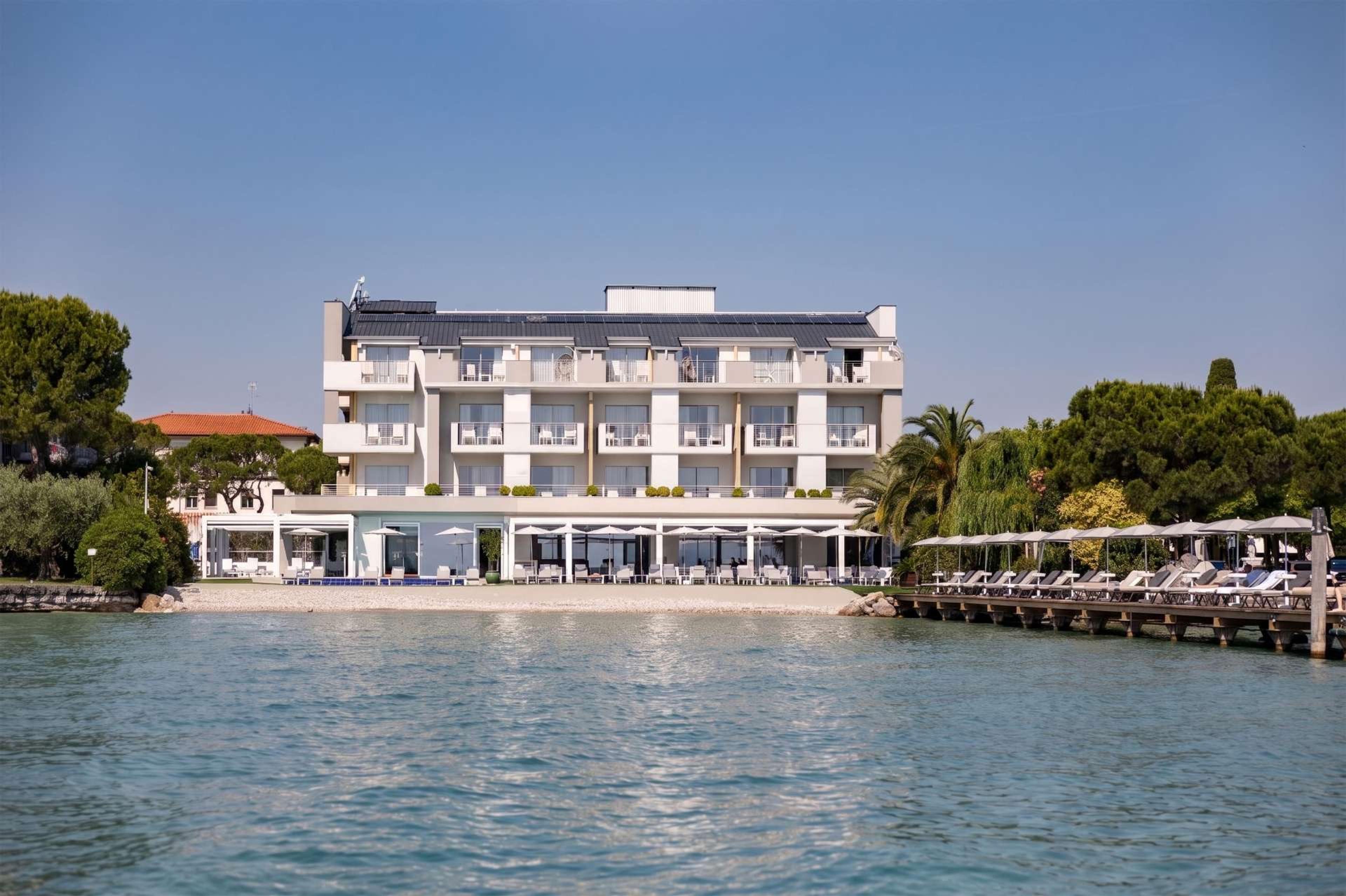 The hotel's white, multi-storey lakefront building seen from the water, with a pool terrace, a beach, and a long dock reaching out over the lake.