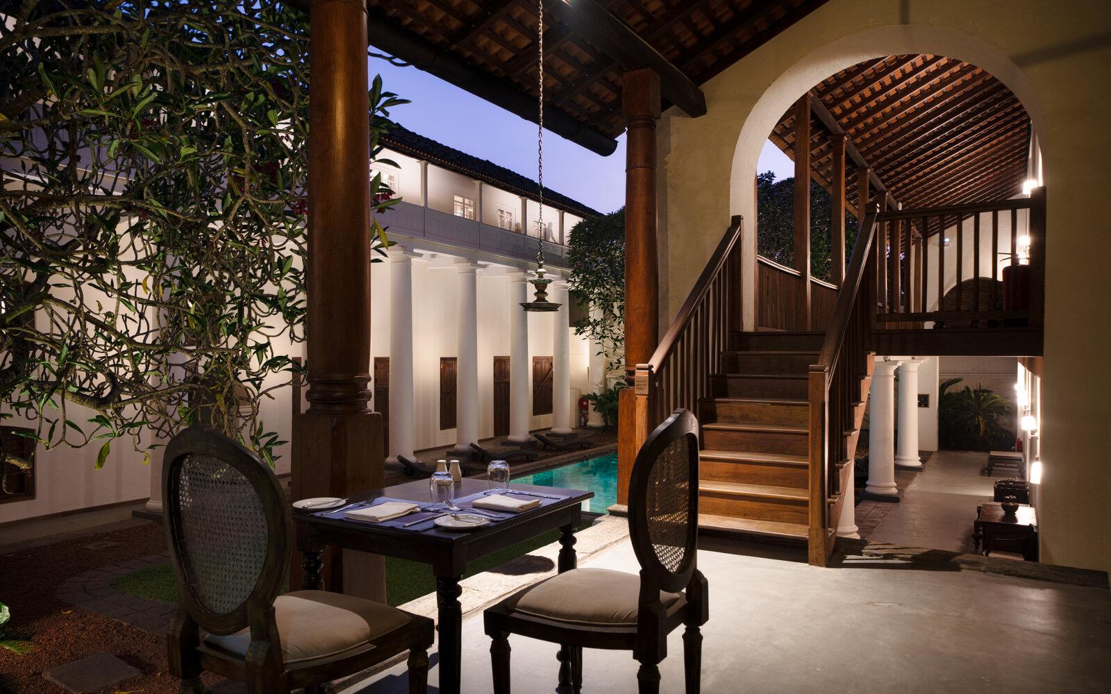 A table set for two beside a pool at dusk, with a wooden staircase and lantern light behind it and the colonnaded building glowing beyond.