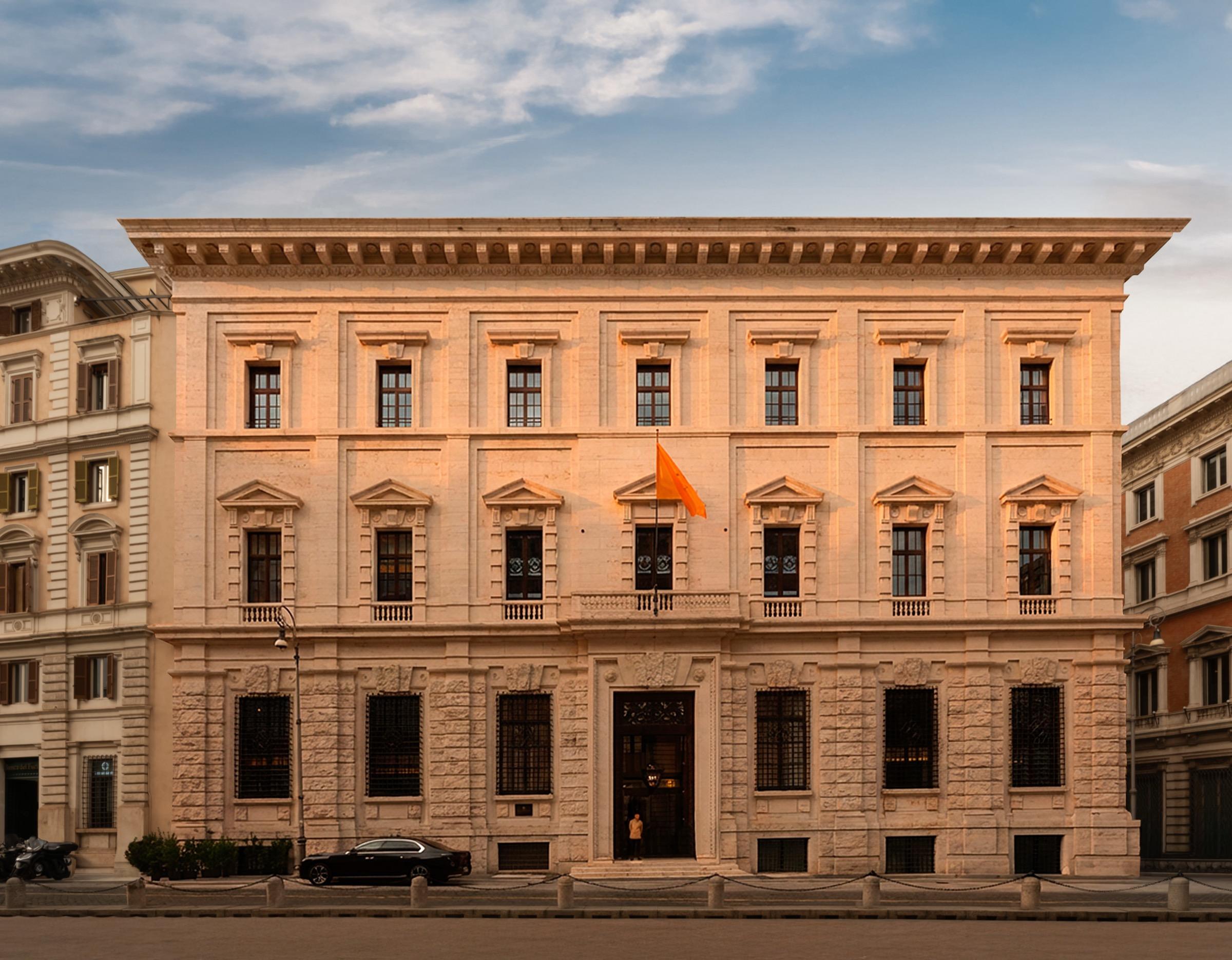 a cream stone palazzo on a Rome street, its windows lit gold by the evening sun, with a car parked by the entrance and a flag above the door