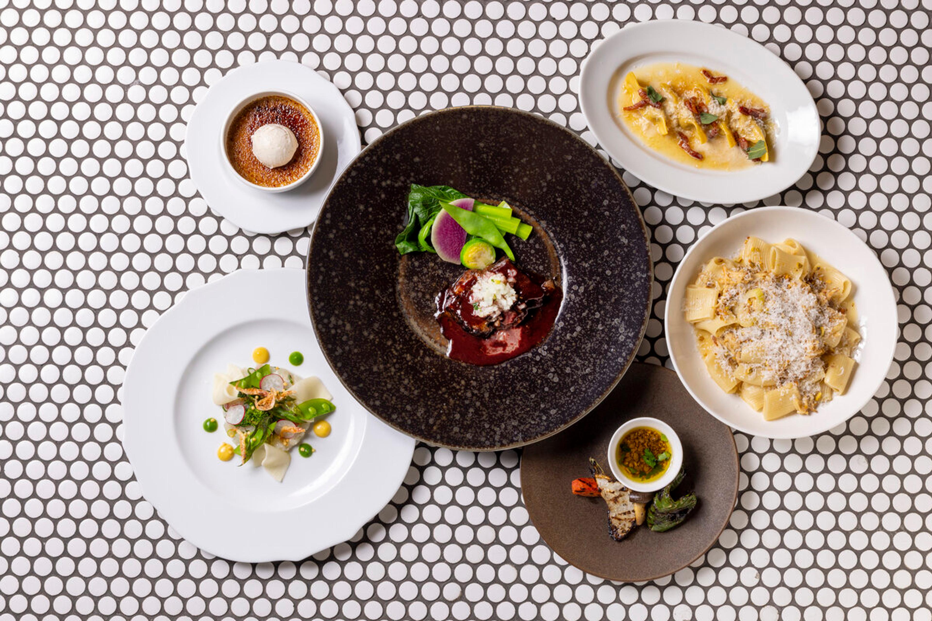 An overhead view of five plated restaurant dishes on a dotted tile surface, including pasta, ravioli, a seared dish in red sauce, a tasting plate and a creme brulee.