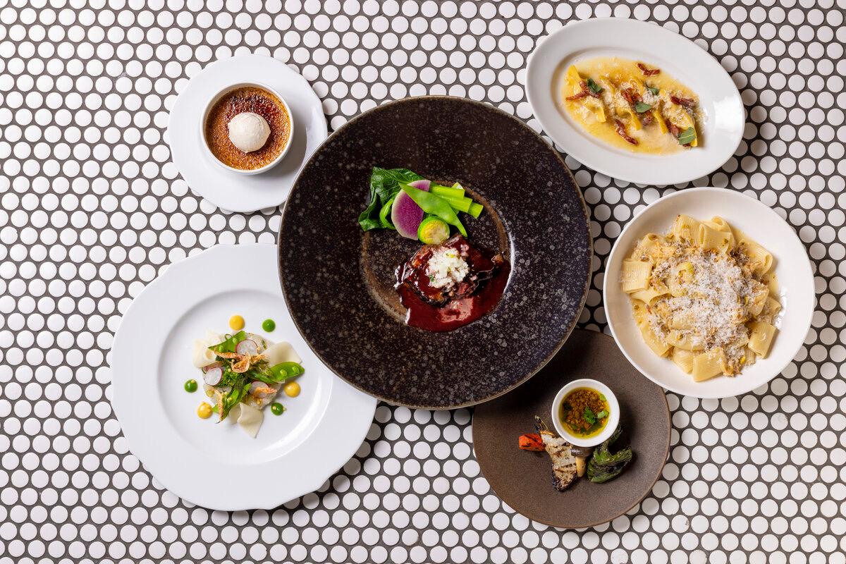 An overhead view of five plated restaurant dishes on a dotted tile surface, including pasta, ravioli, a seared dish in red sauce, a tasting plate and a creme brulee.