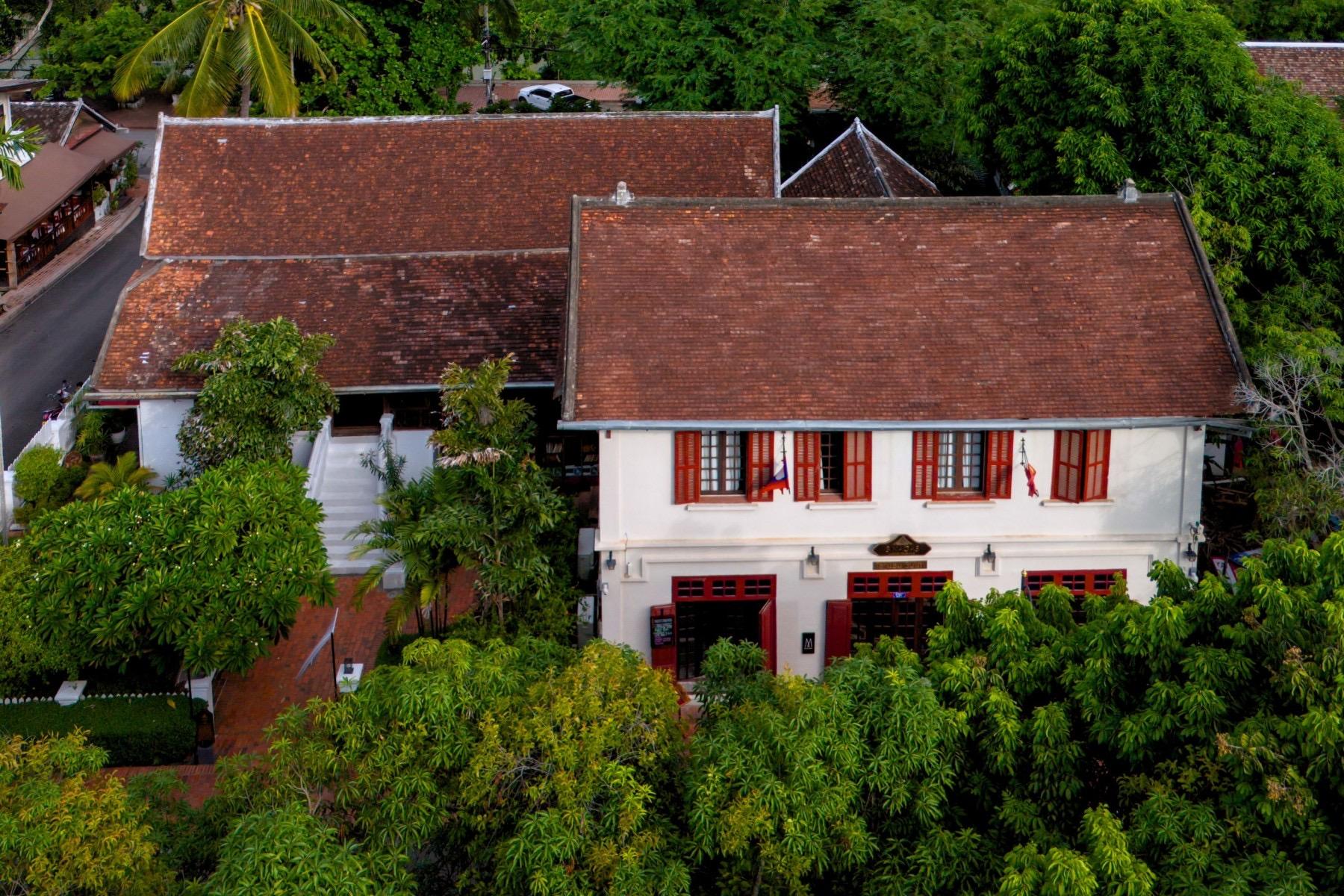 An aerial view of a colonial-style hotel building with red tile roofs and red-shuttered windows, surrounded by mango and palm trees on a quiet street.