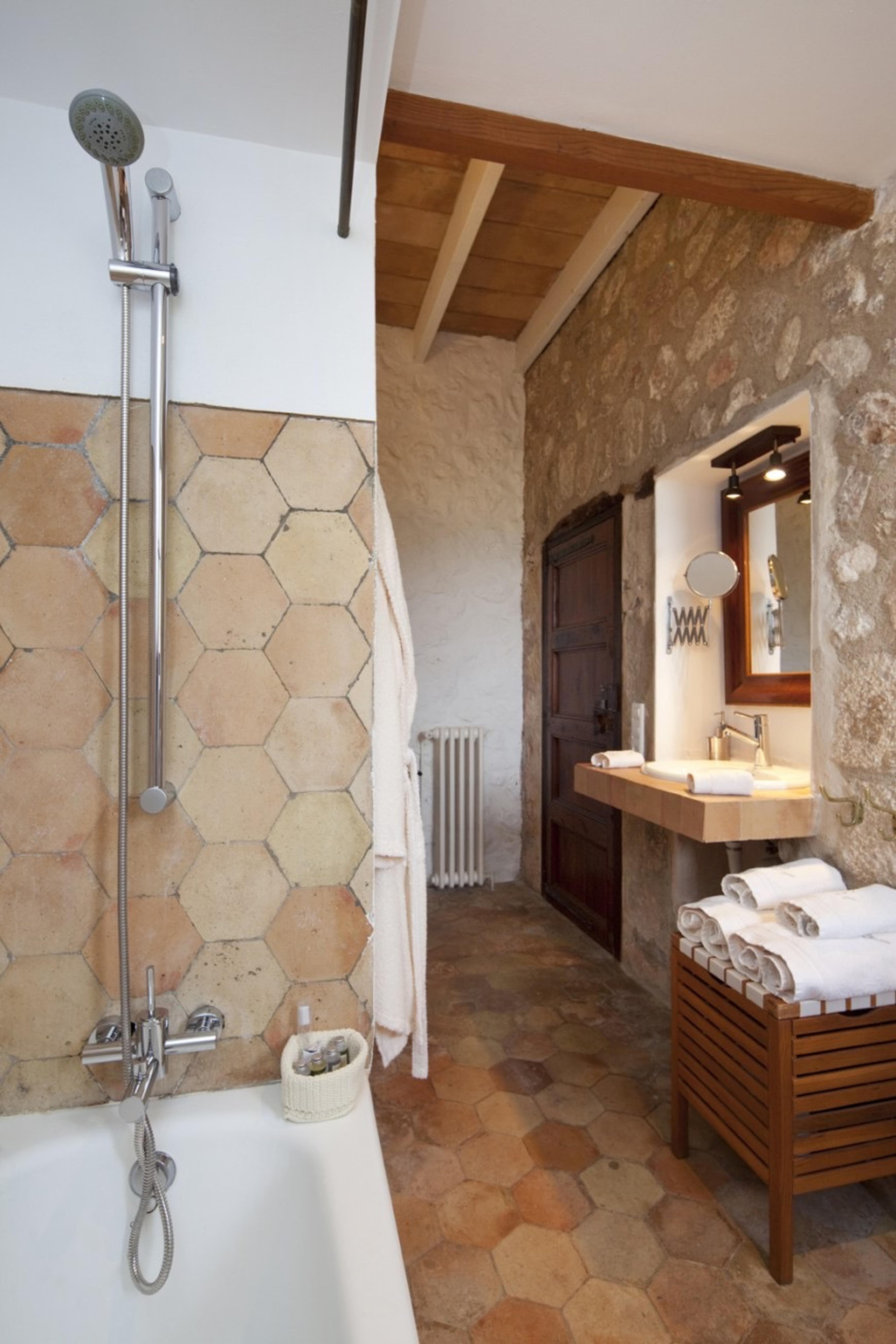 A freestanding bathtub and rain shower on hexagonal tiles, with a stone archway leading to a vanity and towels beyond.