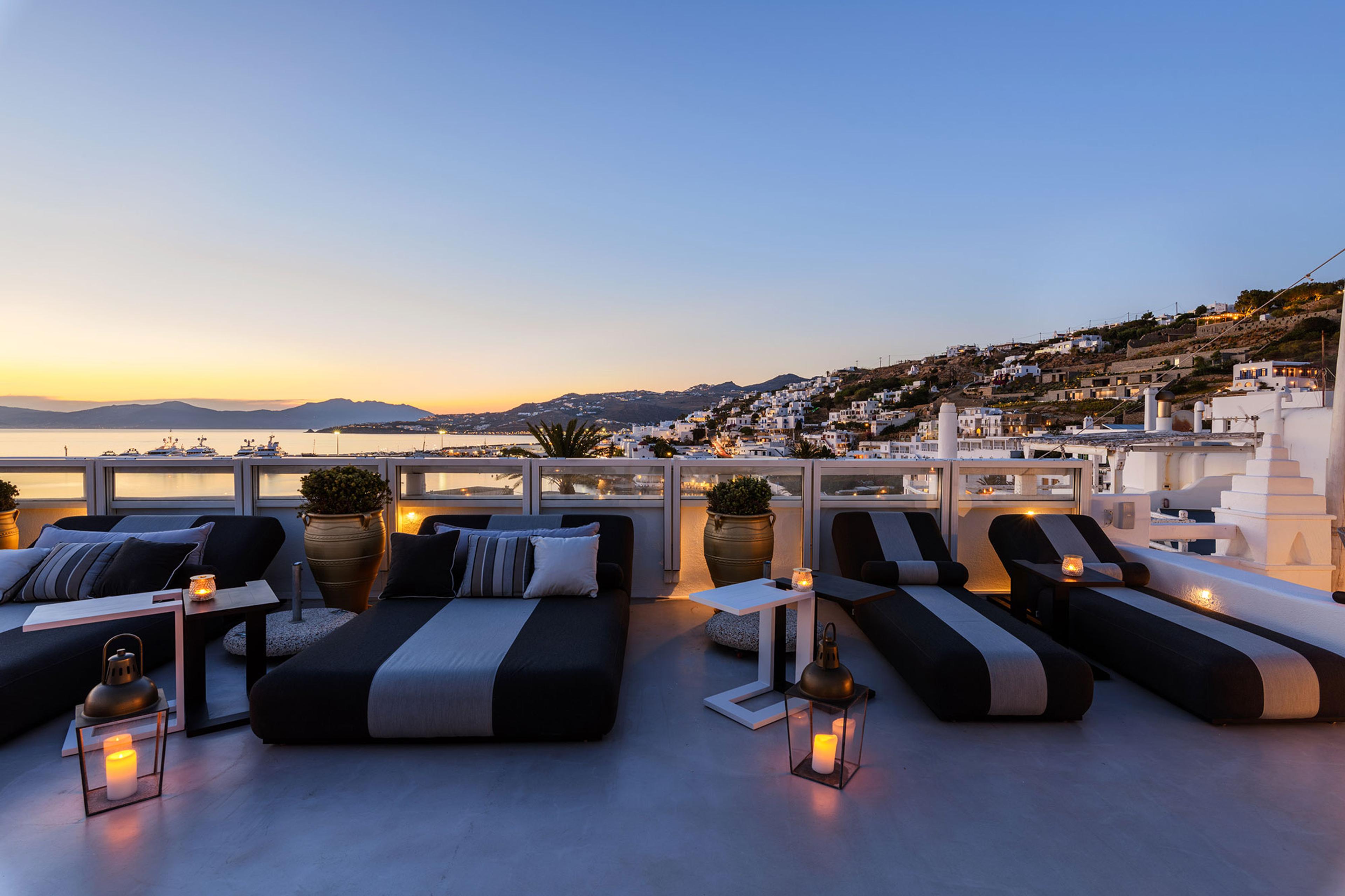 Rooftop daybeds and a sofa overlooking the town and sea at dusk, with lanterns lit on the floor.
