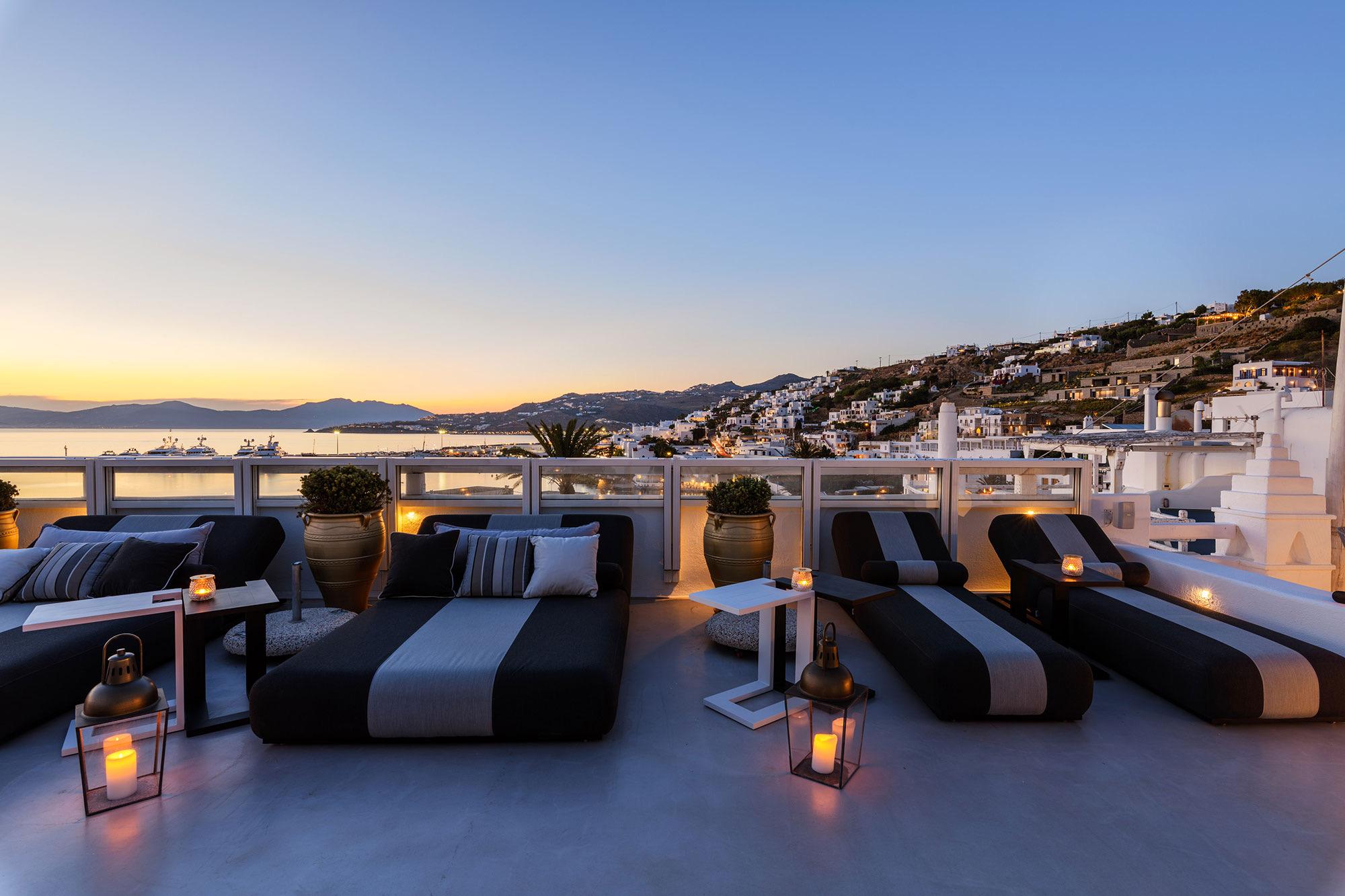Rooftop daybeds and a sofa overlooking the town and sea at dusk, with lanterns lit on the floor.