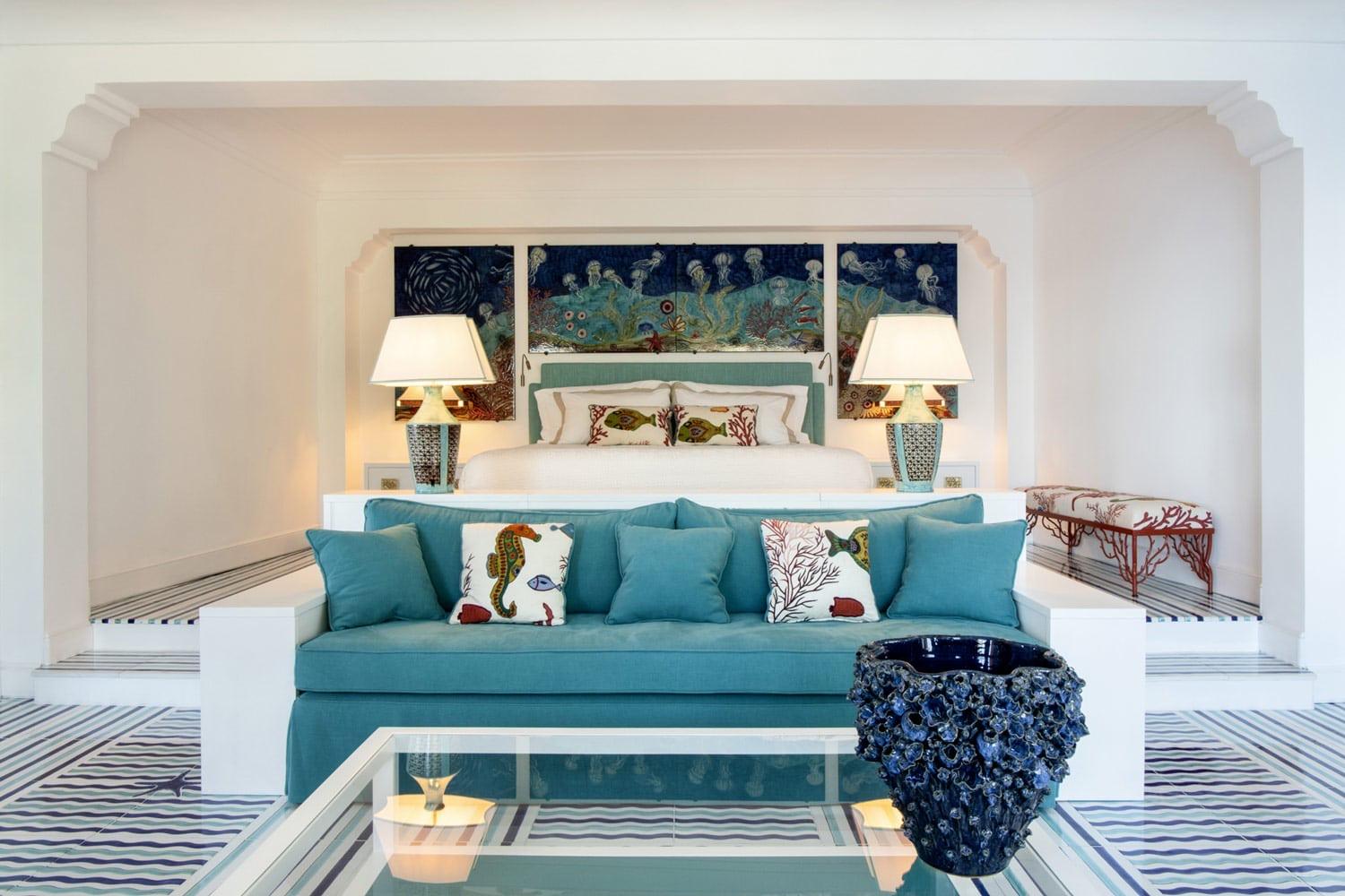 A hotel suite living room with a teal sofa covered in sea-themed cushions, a glazed blue vase, and artwork of jellyfish above the headboard.
