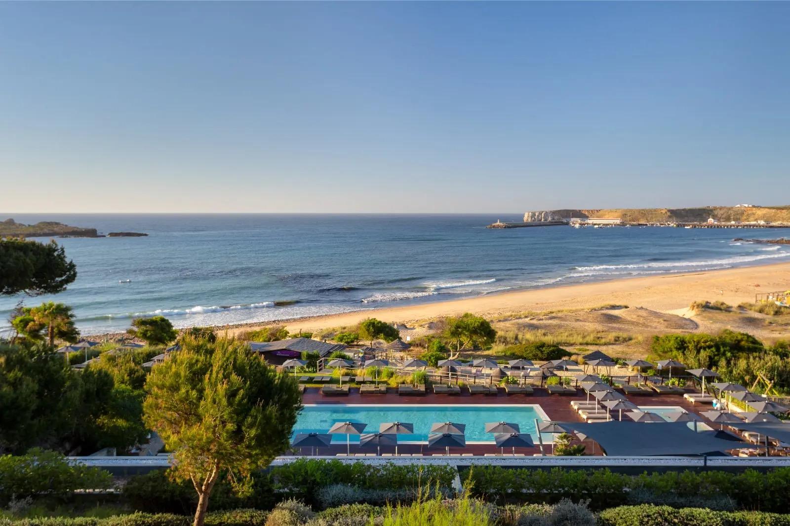An aerial view of a pool with grey parasols, a grassy slope and a sandy beach, with the sea and a headland beyond.