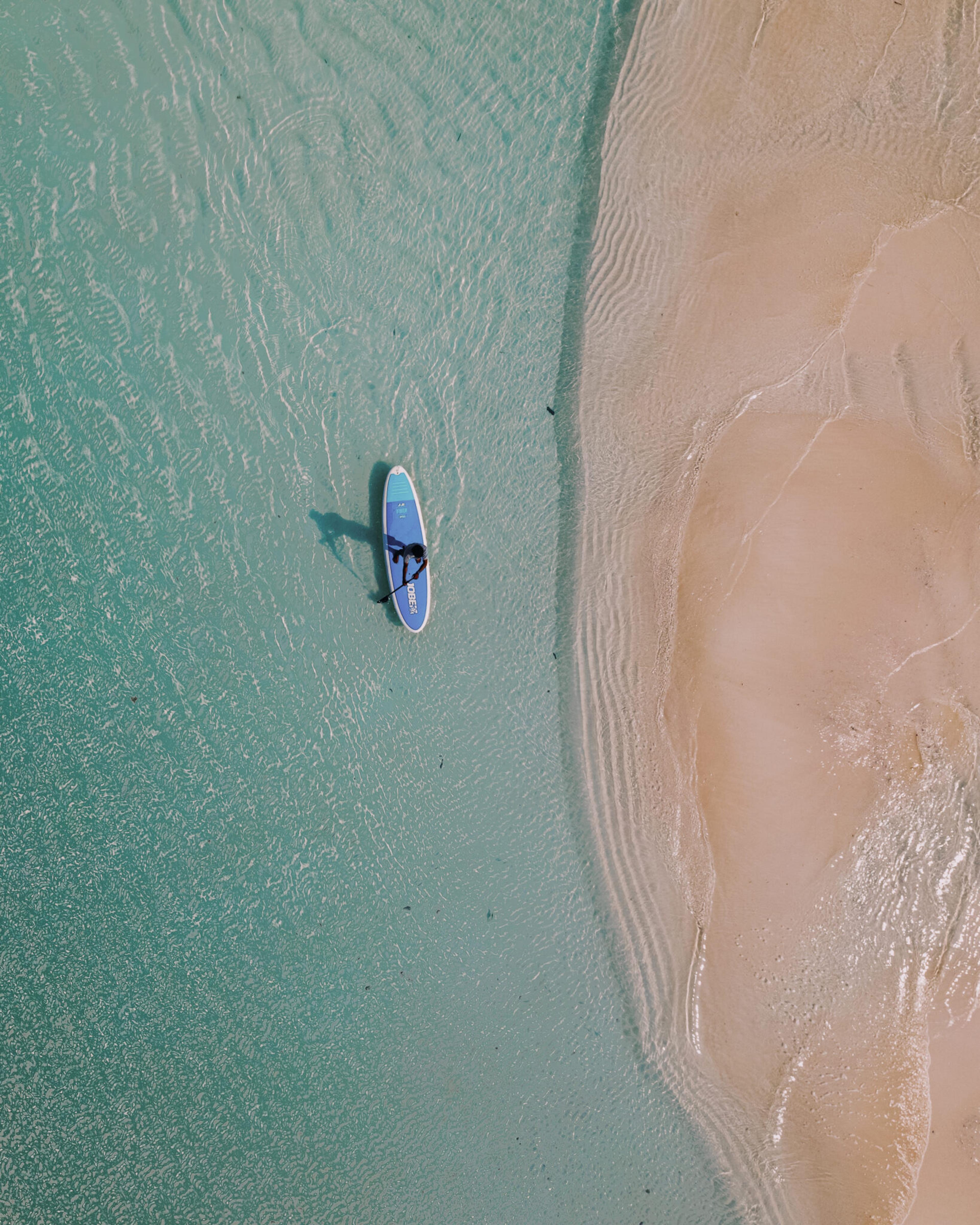 a person on a paddleboard in shallow turquoise water beside a sandbar, seen from directly above