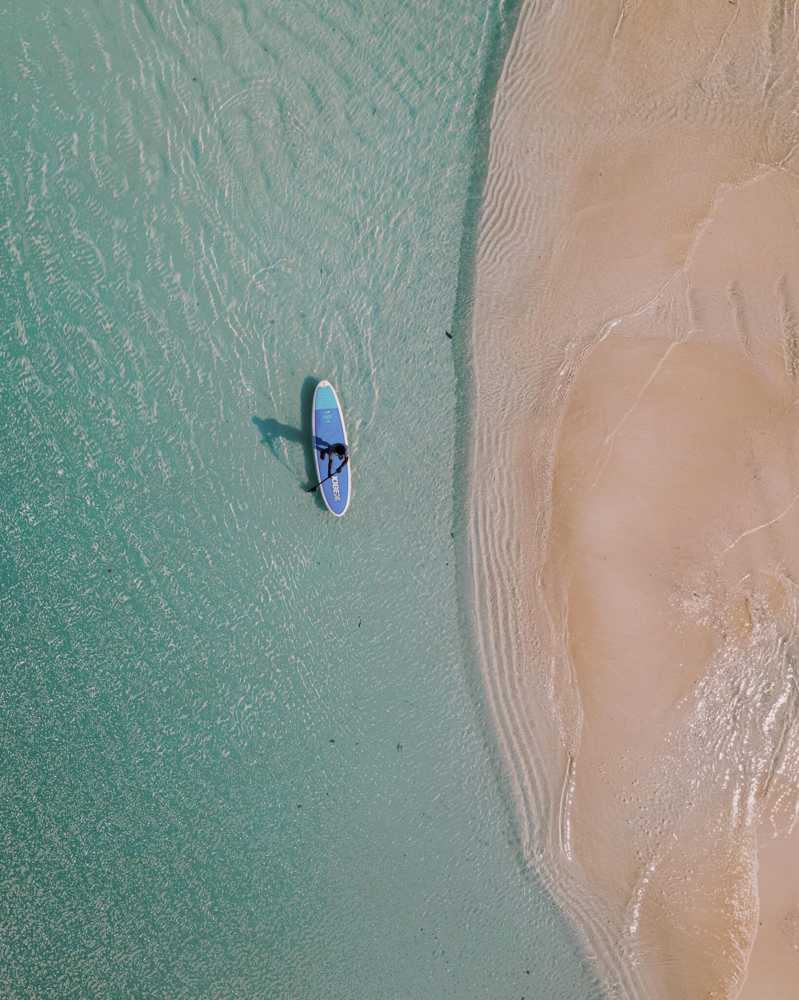 a person on a paddleboard in shallow turquoise water beside a sandbar, seen from directly above