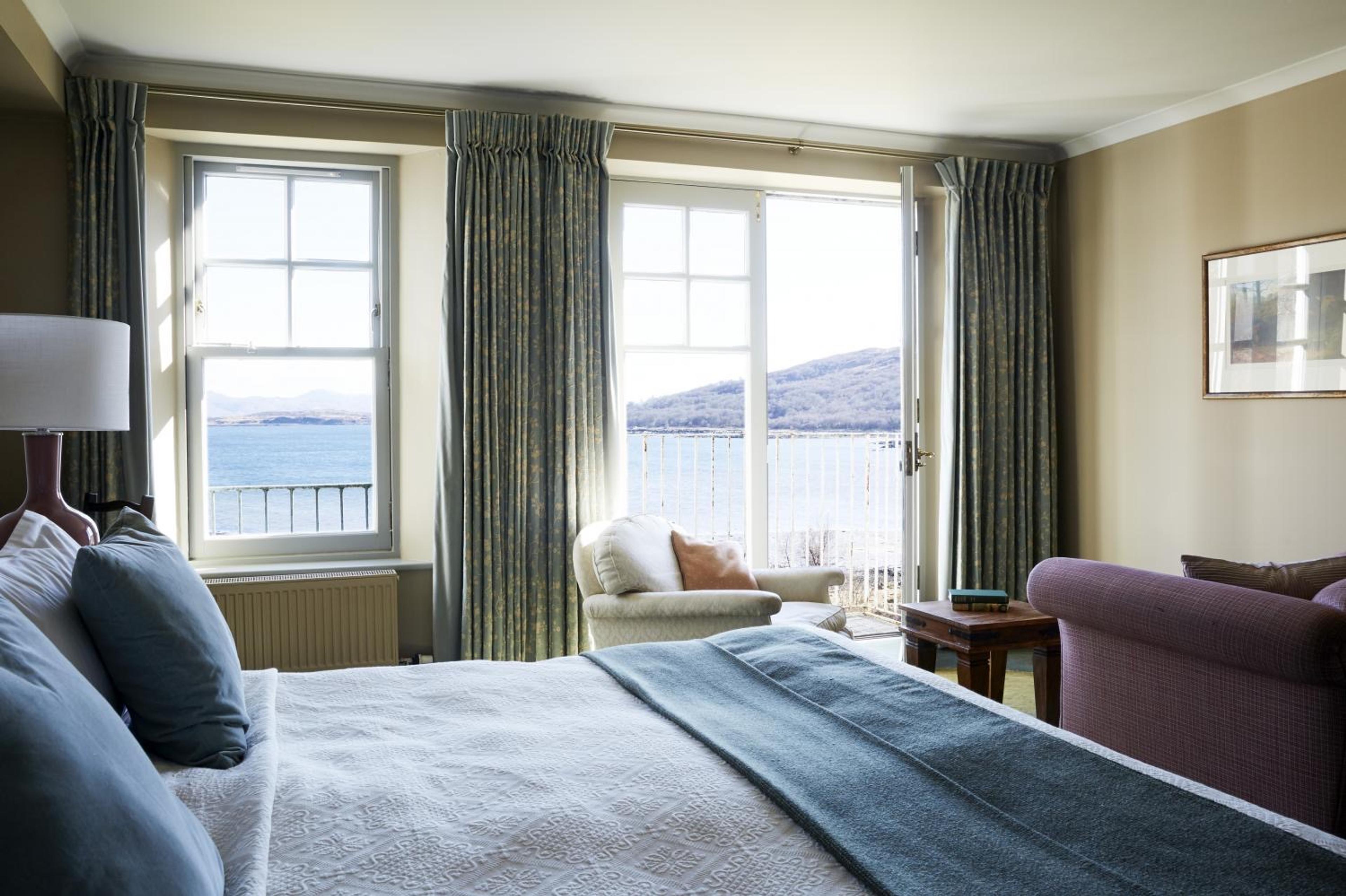 A bedroom with a white bed and an armchair in front of tall curtained windows open to a balcony overlooking a sea loch and hills.