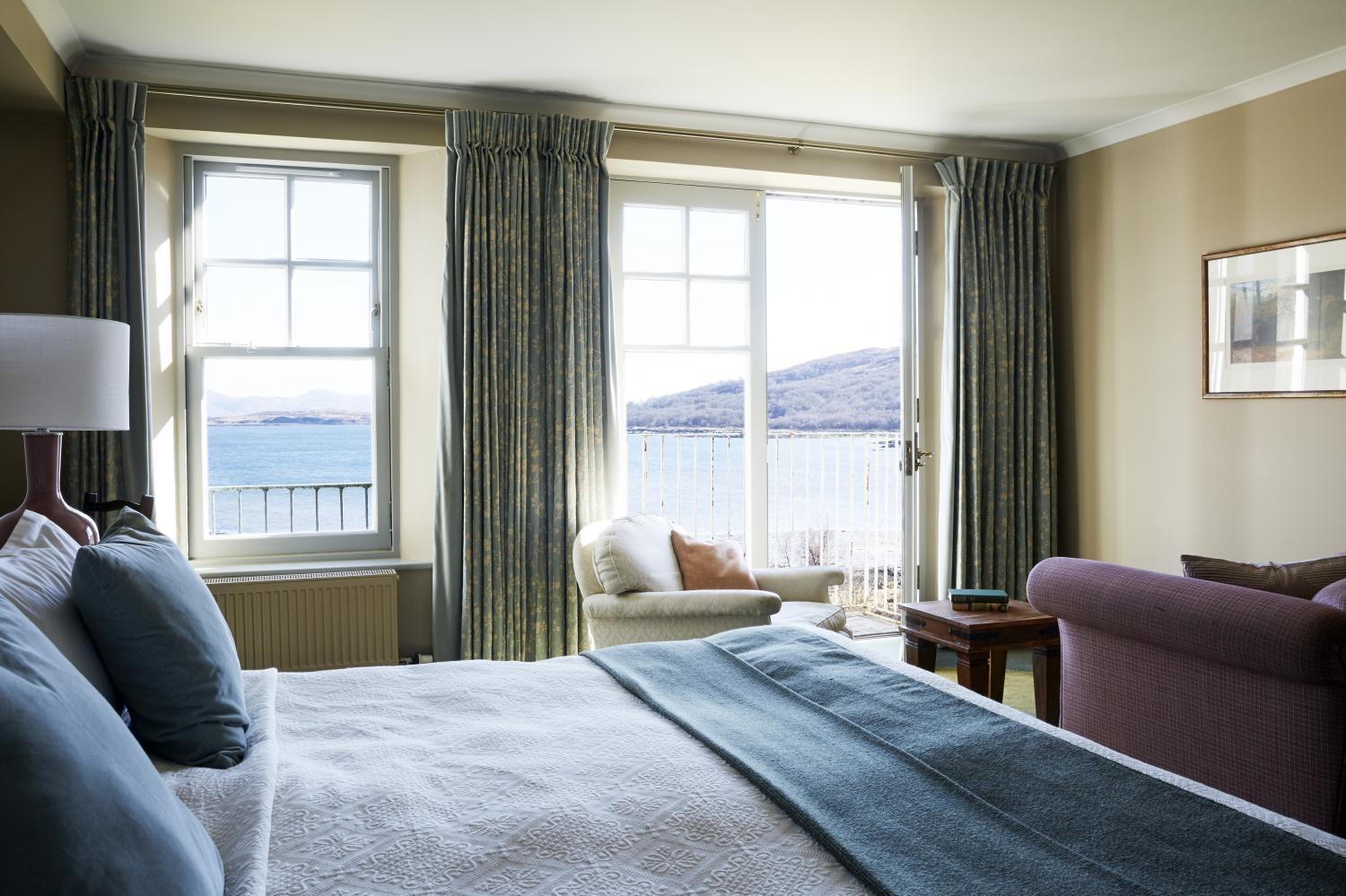 A bedroom with a white bed and an armchair in front of tall curtained windows open to a balcony overlooking a sea loch and hills.