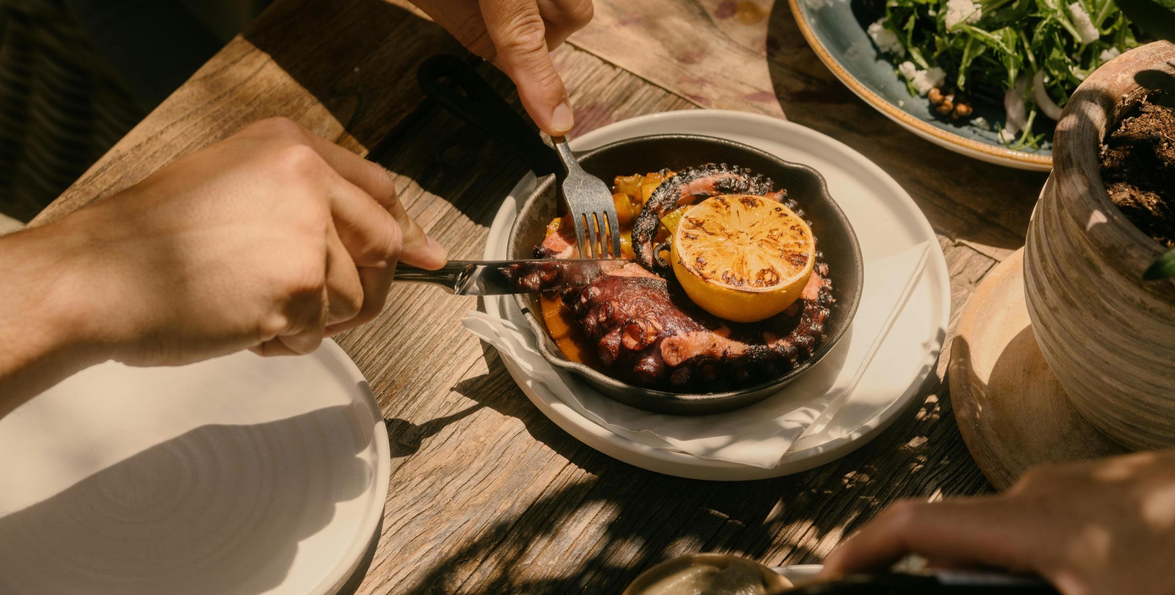 A hand cutting into a dish of grilled octopus with charred lemon, next to a green salad on a wooden table.