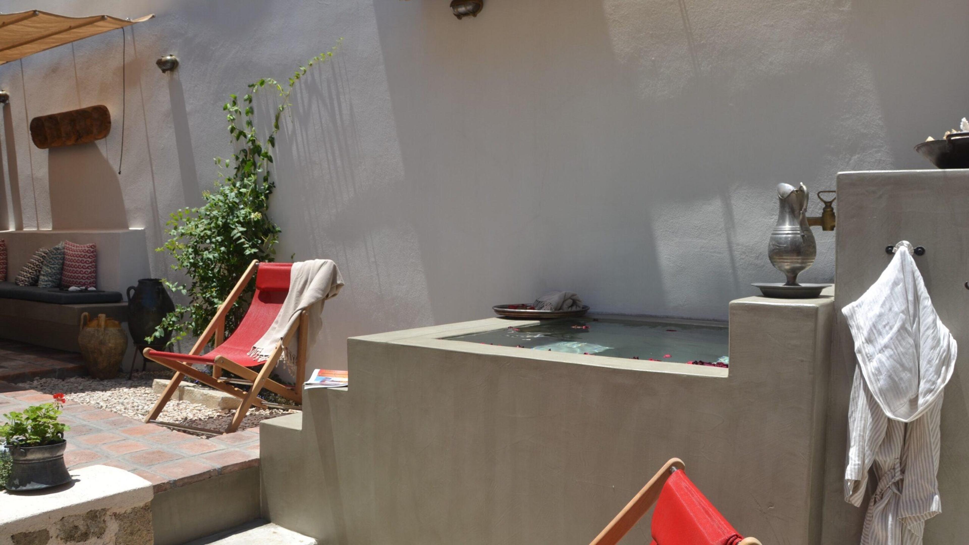 An outdoor plunge pool in sunlight, with a red deck chair and a robe hanging nearby.