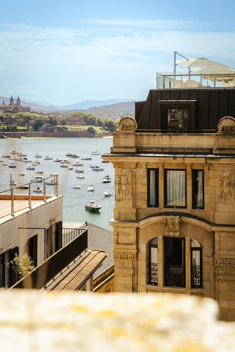 A hotel's corner tower and facade seen from above, with a rooftop terrace and umbrella on top, a bay full of small boats, and hills beyond.