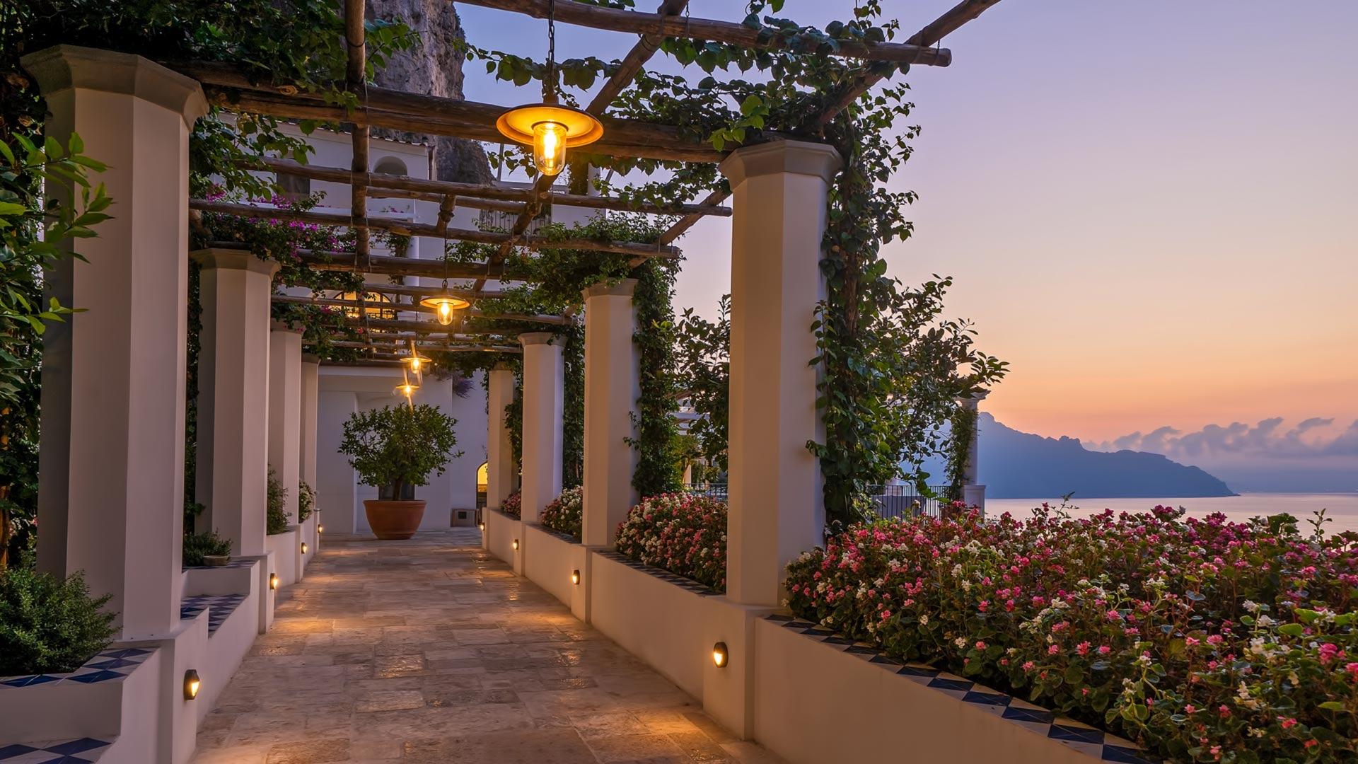 A lit pergola walkway with hanging lanterns and flowering shrubs along one side, leading past a potted tree toward the hotel at dusk, the sea and hills beyond lit by the sunset.