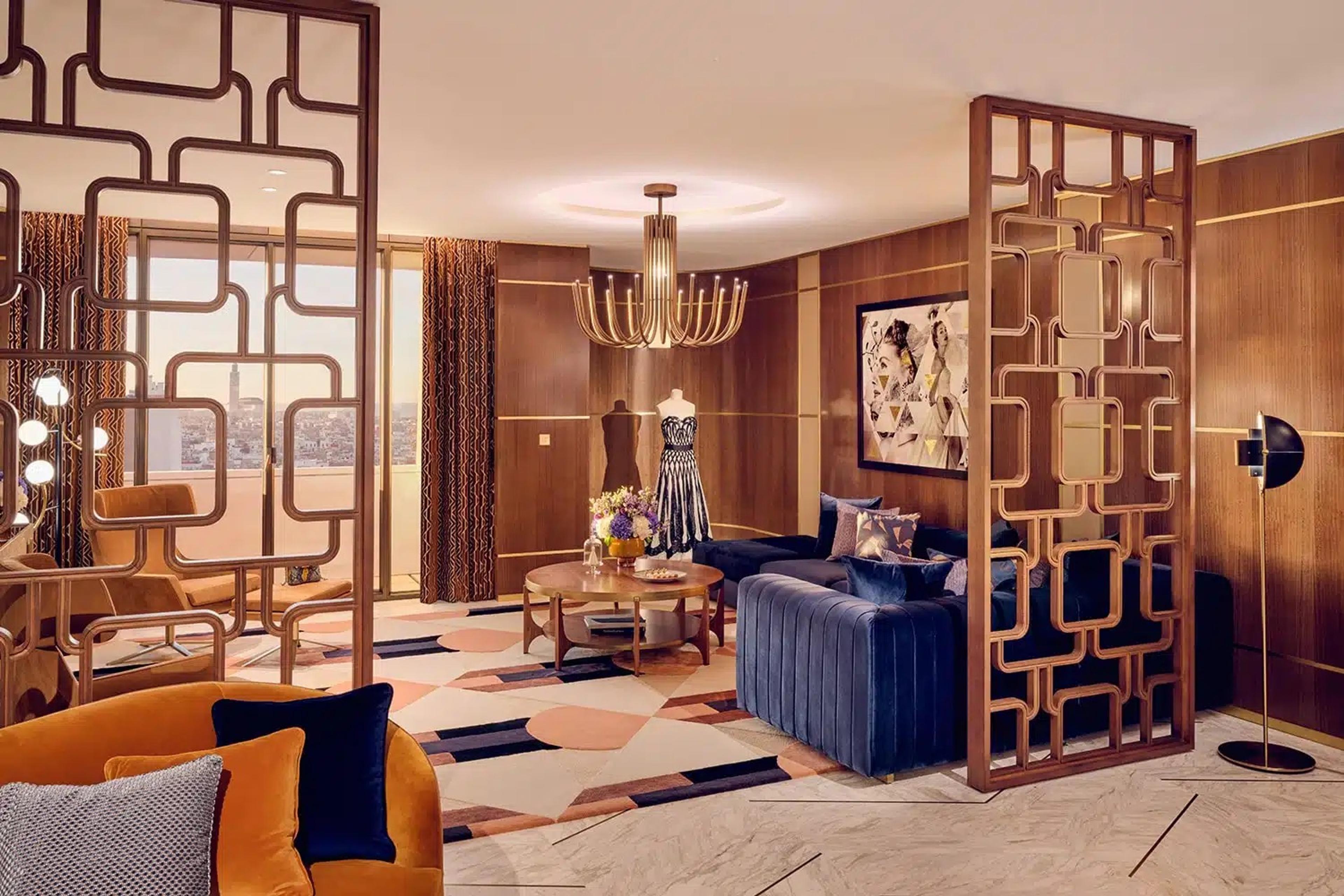 A hotel suite living room with a blue sofa, orange armchair, patterned screens and a view over rooftops through tall windows.