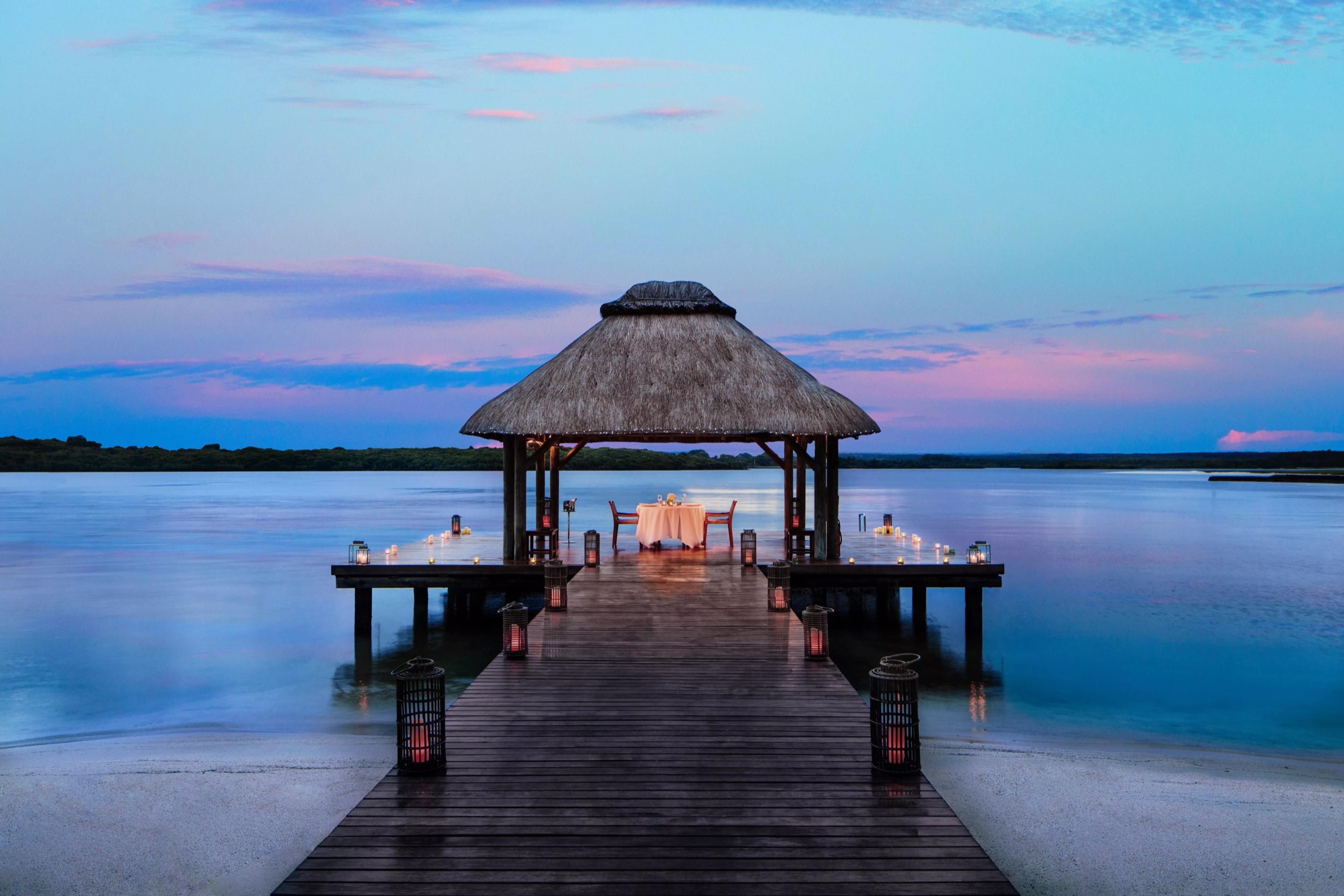 A thatched-roof pavilion at the end of a wooden jetty holds a table set for two, lit by lanterns at dusk with the sea on both sides.