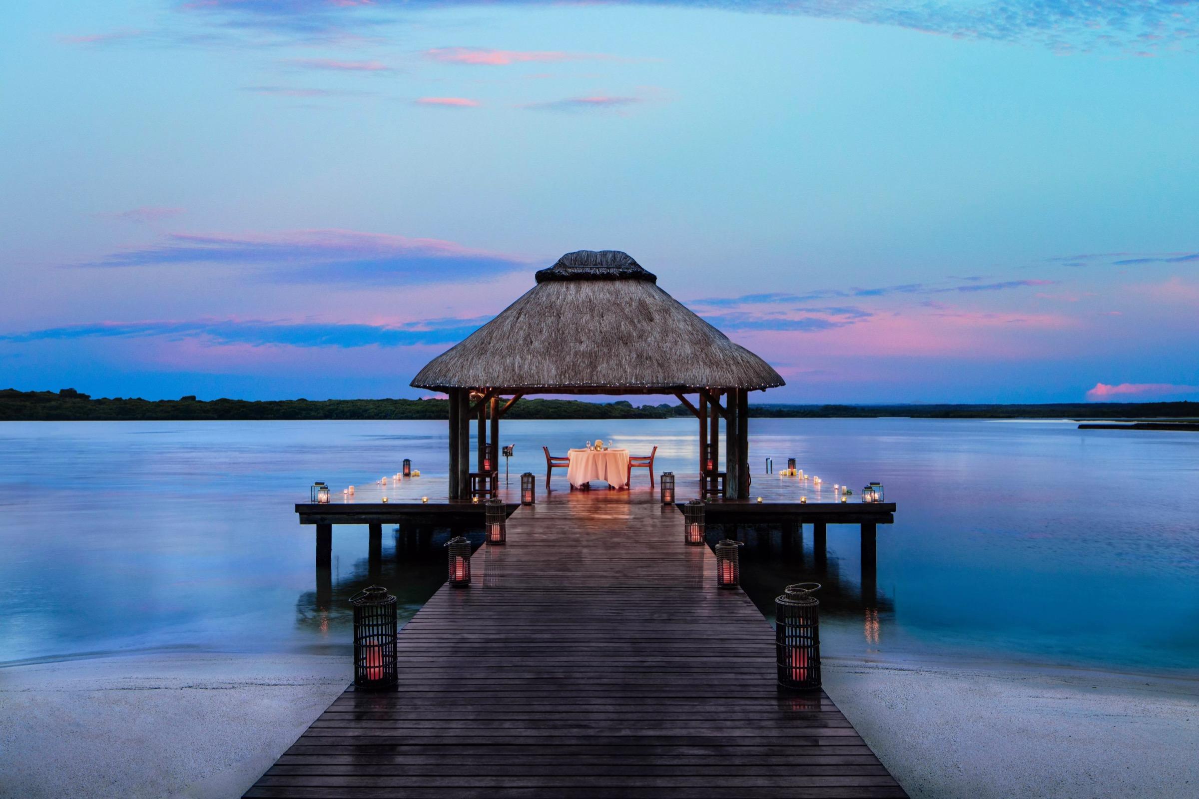 A thatched-roof pavilion at the end of a wooden jetty holds a table set for two, lit by lanterns at dusk with the sea on both sides.