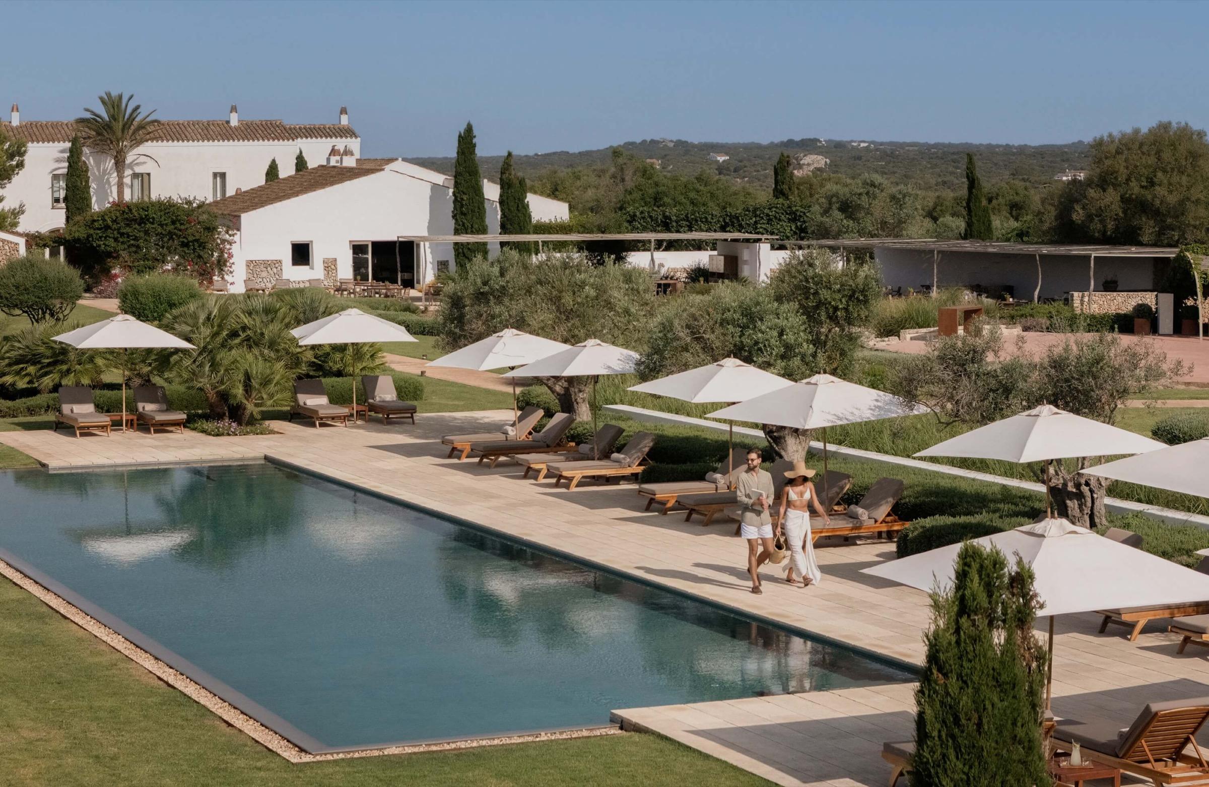 Two people walk past a long swimming pool lined with sun loungers and umbrellas, with the main white building and hills behind.