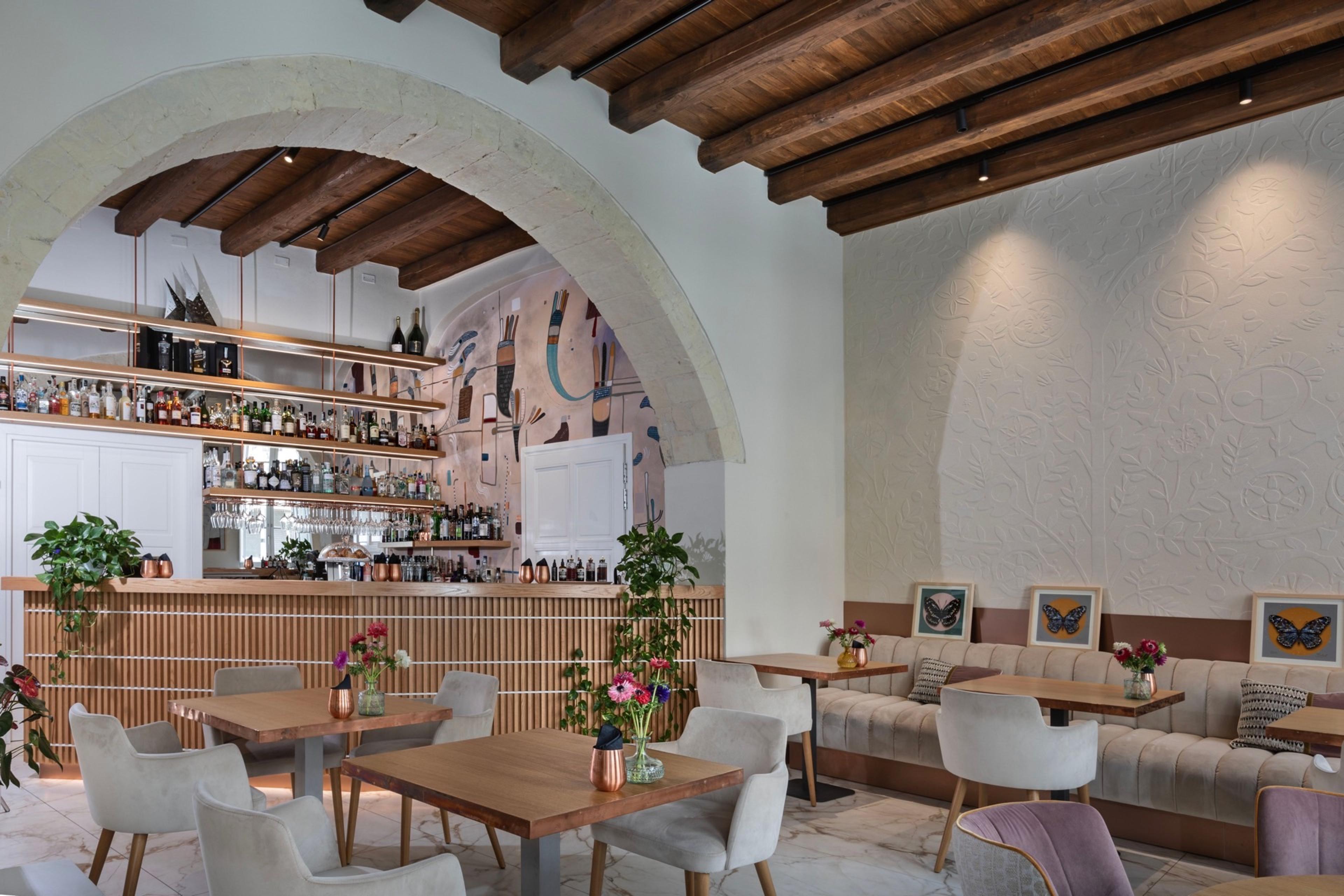 A bar and dining area under a stone arch, with wooden shelves of bottles, marble-topped tables and chairs, and framed butterfly prints on a textured wall.