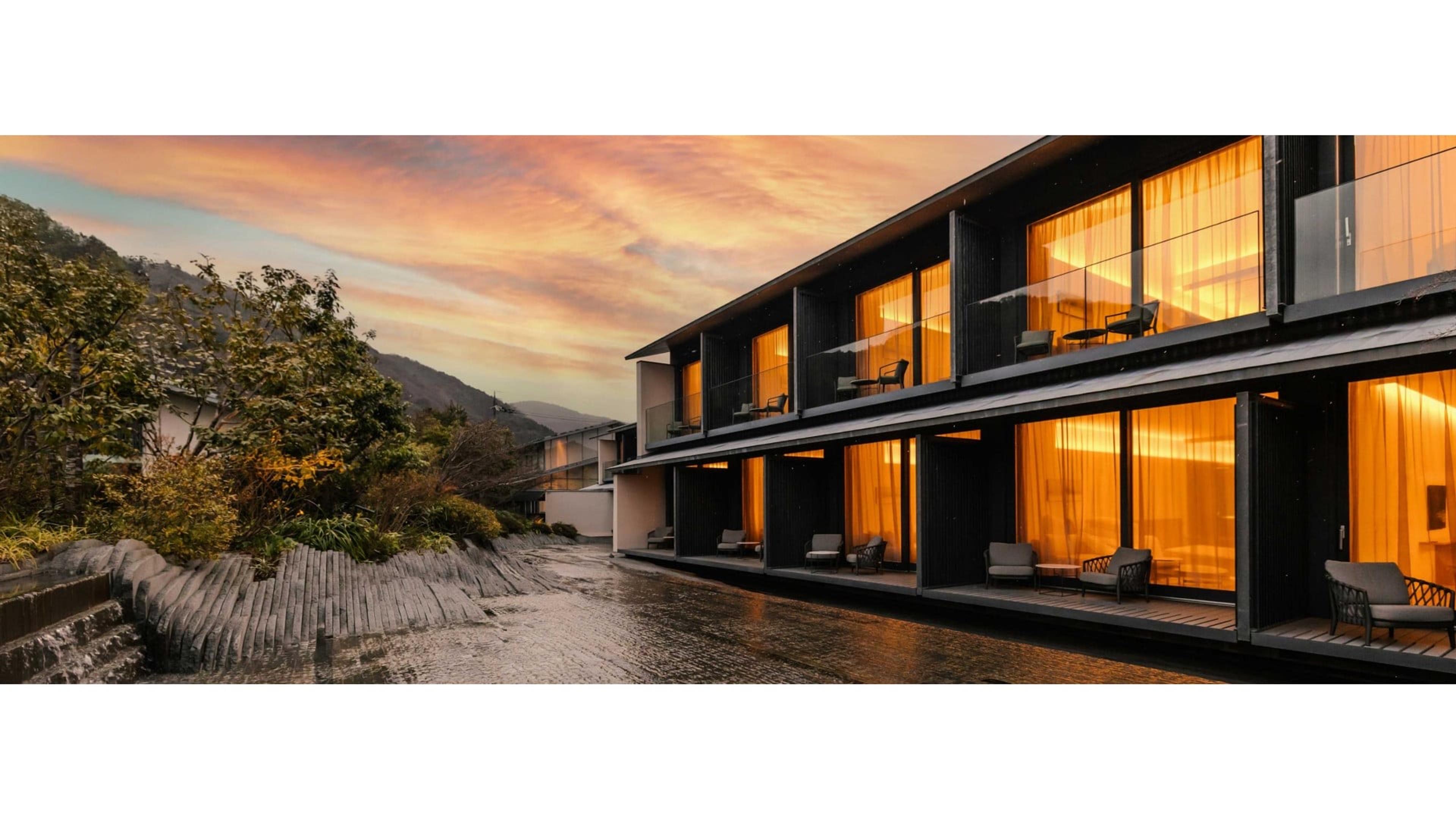 A hotel building at sunset, its glass-fronted rooms lit from within, overlooking a stone riverbed with wooded hills behind