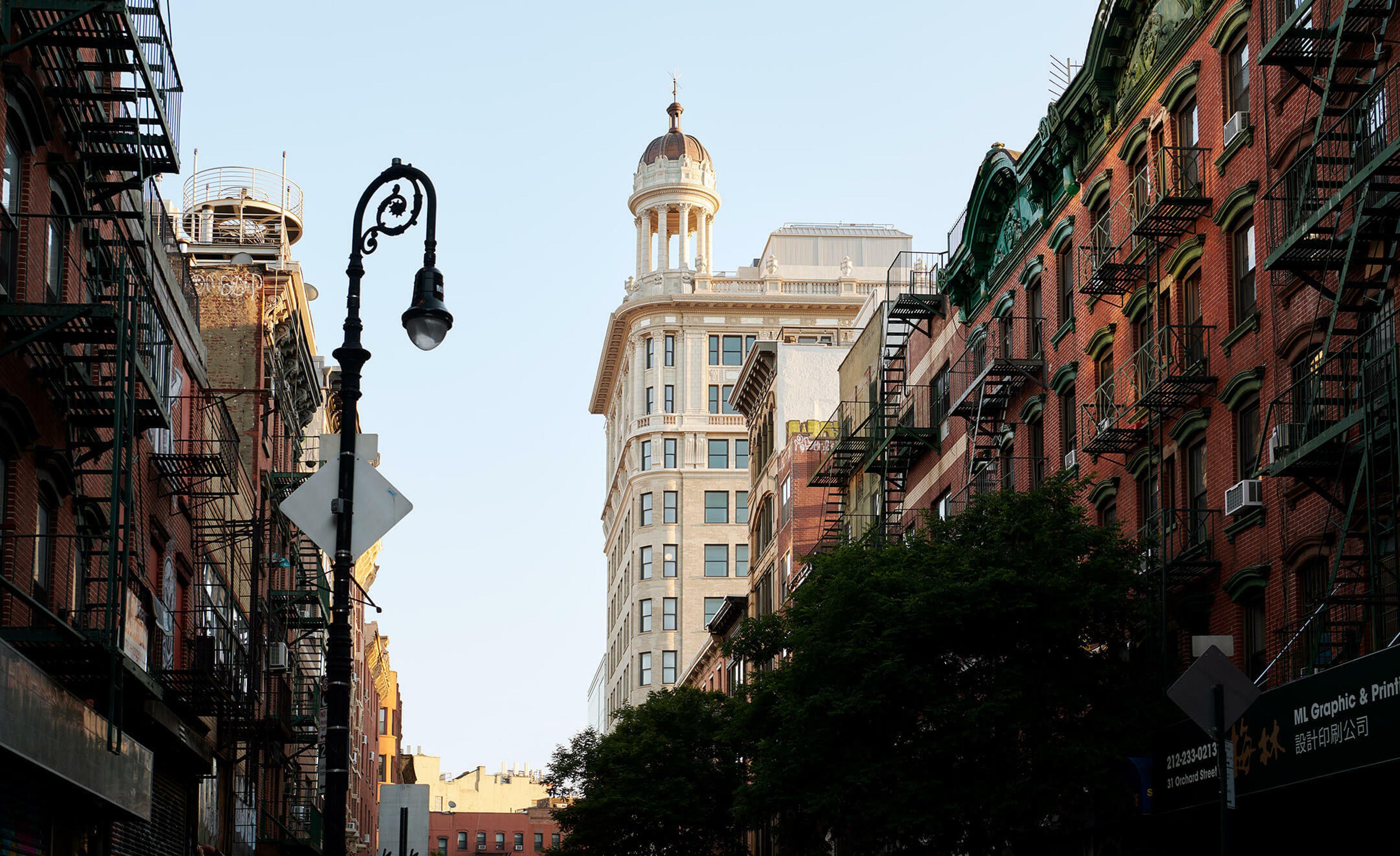 A street-level view of a tiered stone tower with a domed cupola, seen between brick buildings with fire escapes, under a street lamp.