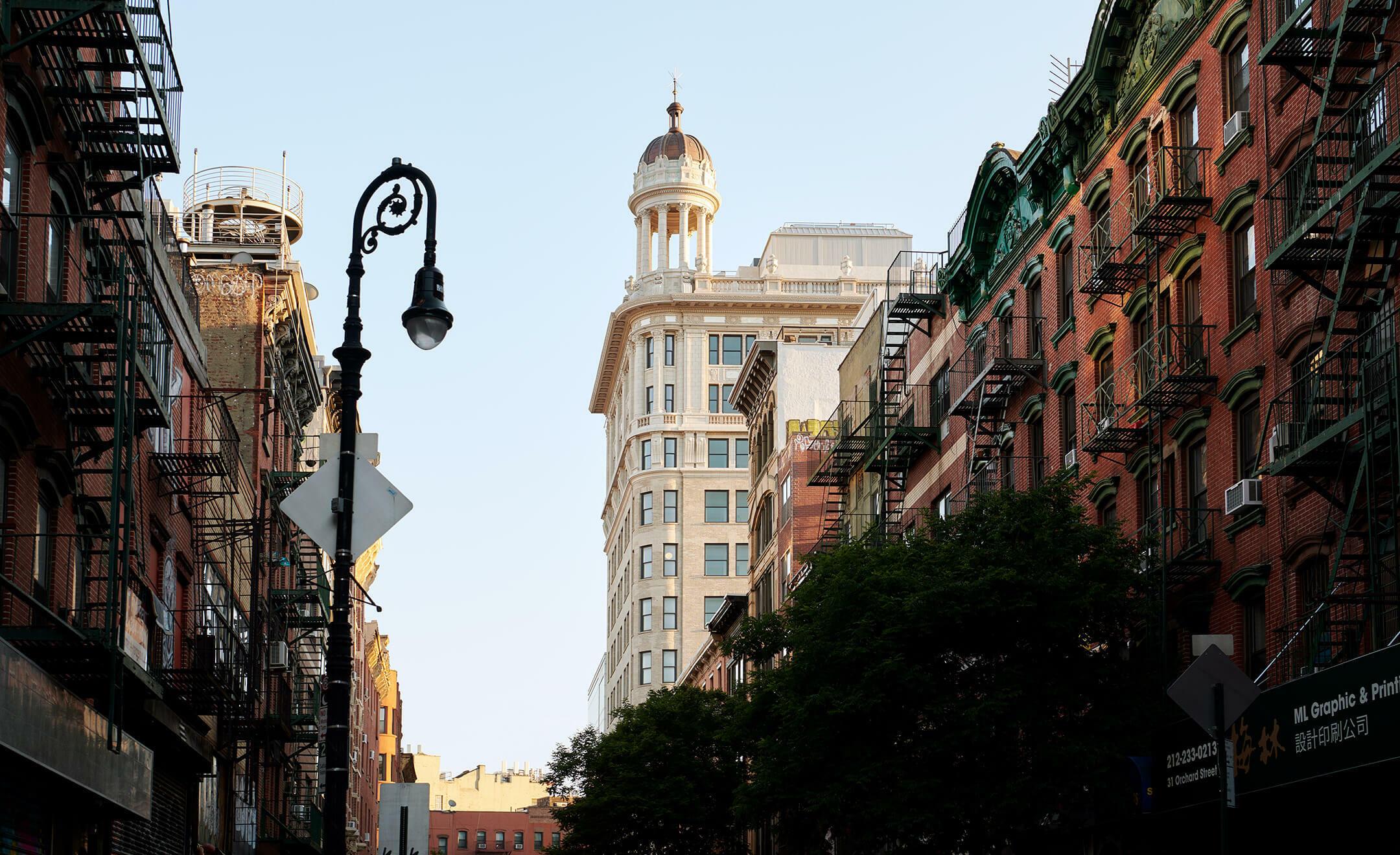A street-level view of a tiered stone tower with a domed cupola, seen between brick buildings with fire escapes, under a street lamp.