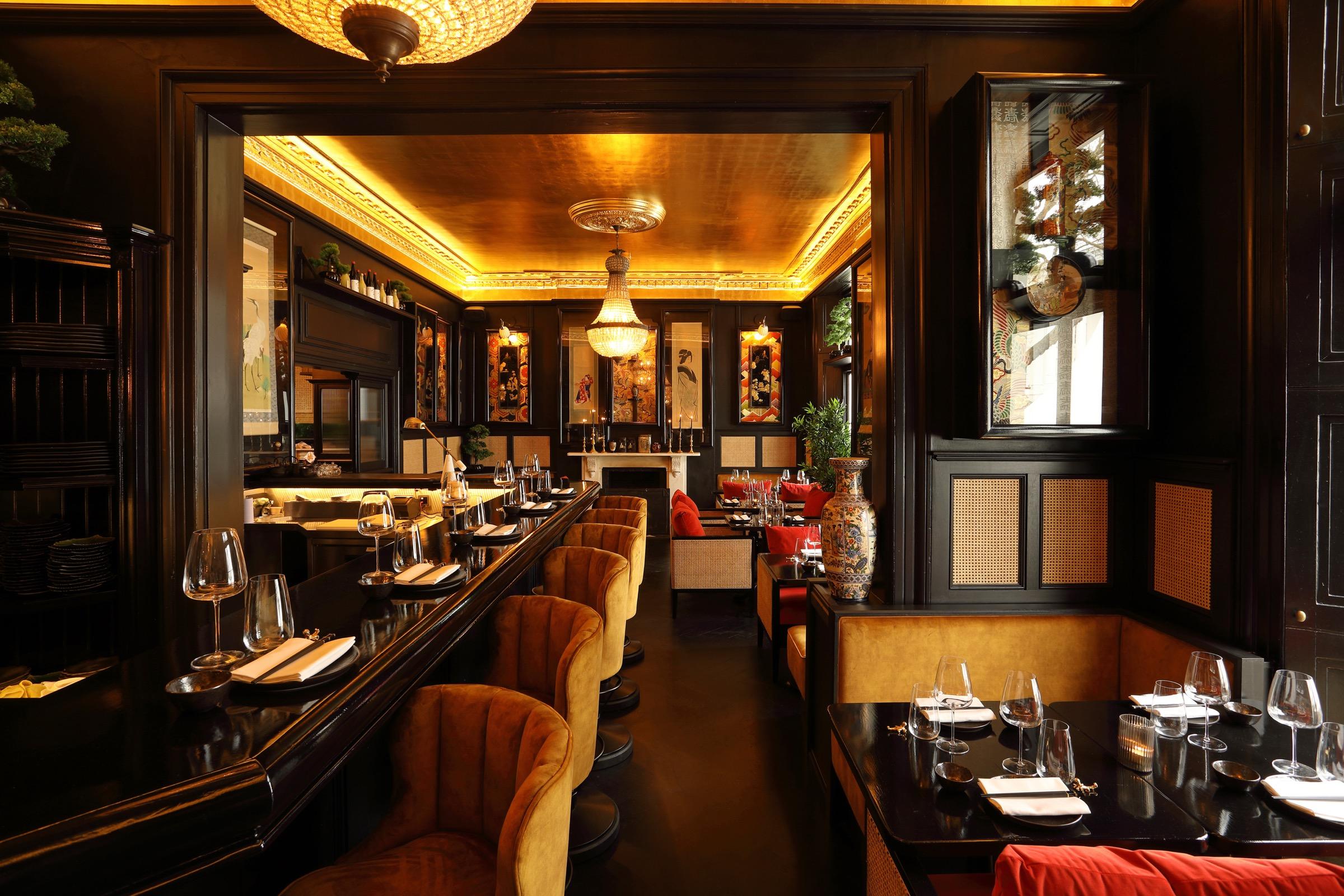 A dimly lit bar and dining room with a gold ceiling, a long velvet-stooled bar, booths and hanging art, seen through an open doorway.