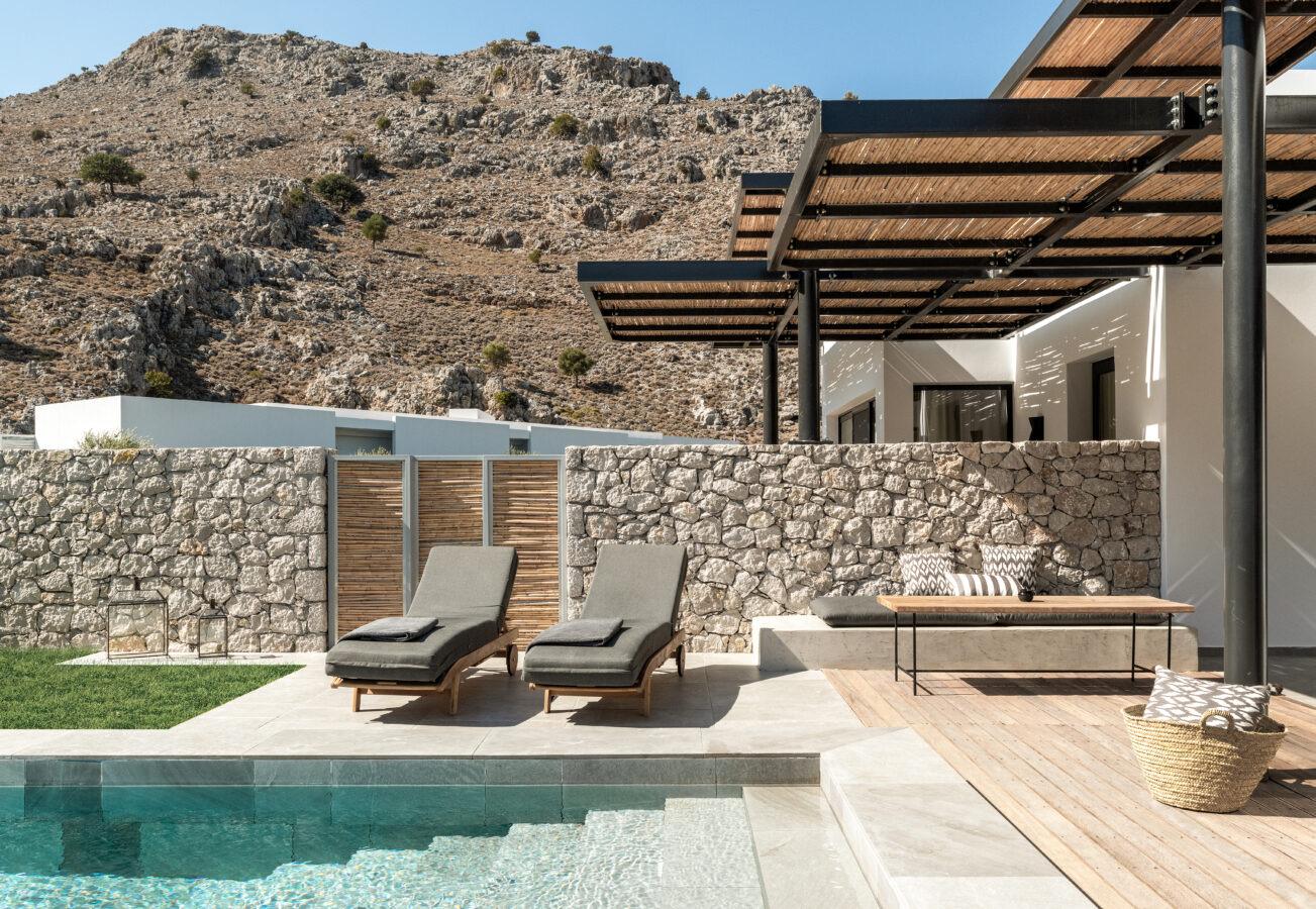 A private plunge pool sits in front of a stone wall, with two grey loungers on a pale terrace, and a bench under a reed-roofed pergola on timber decking with a dry hillside above.