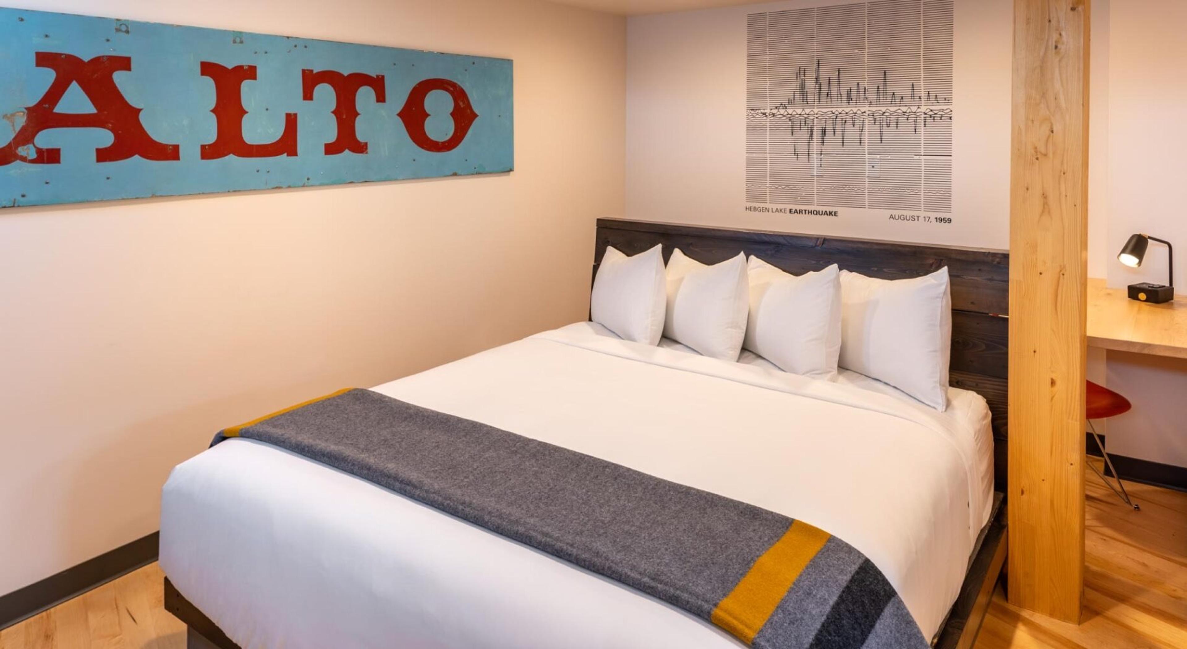 A king bed with white bedding and a grey and yellow throw, under a turquoise sign reading ALTO, with a wooden post and a desk beside it.