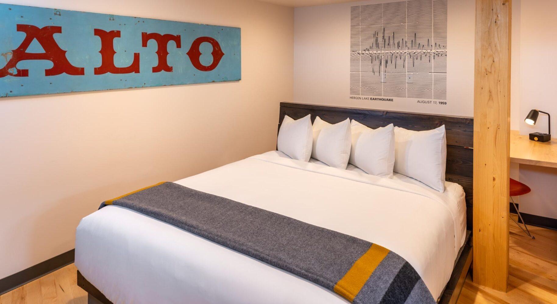 A king bed with white bedding and a grey and yellow throw, under a turquoise sign reading ALTO, with a wooden post and a desk beside it.
