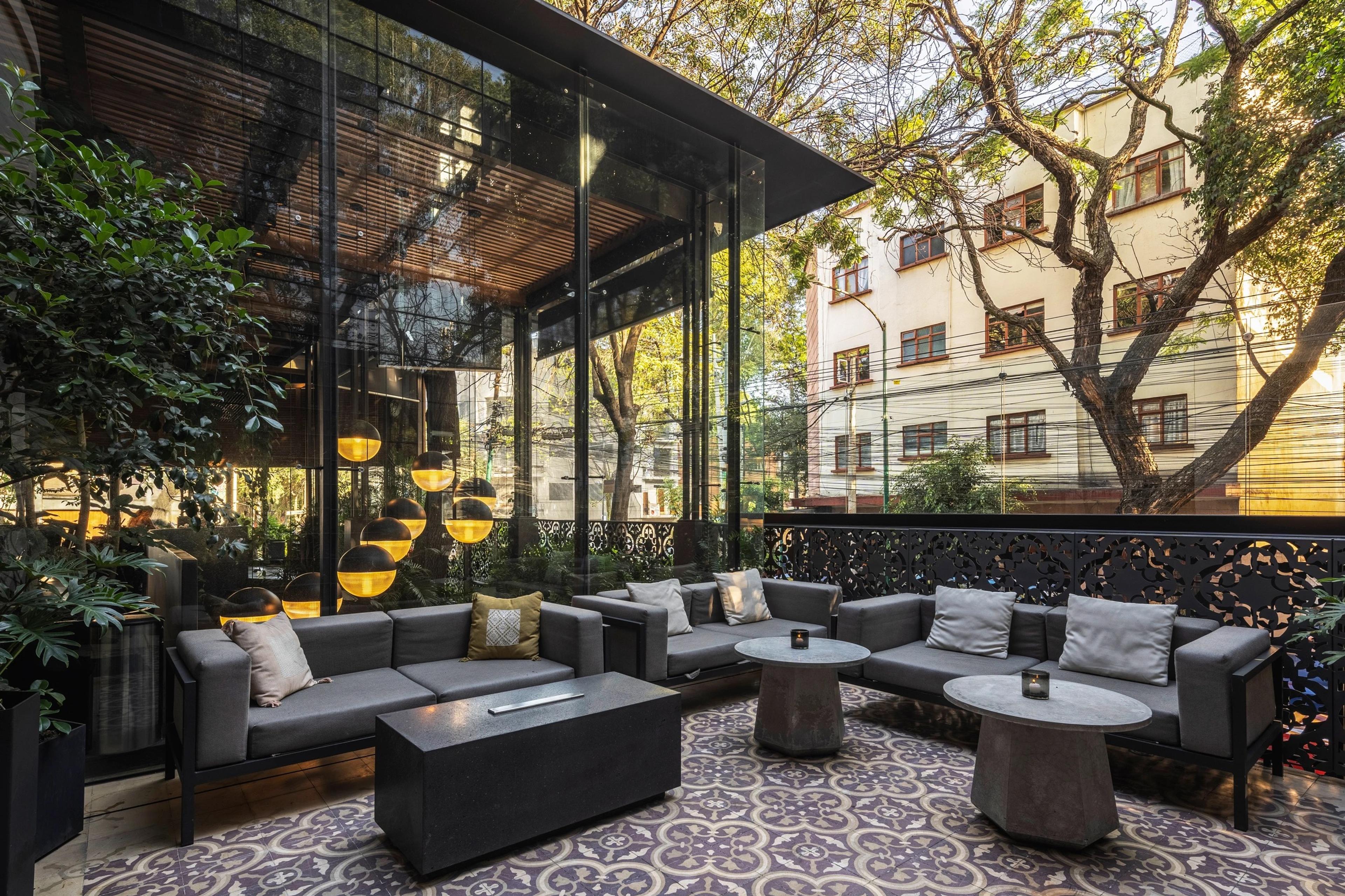 An outdoor lounge with grey sofas and orange cushions on a patterned tile floor, glass walls reflecting trees and a residential street beyond.
