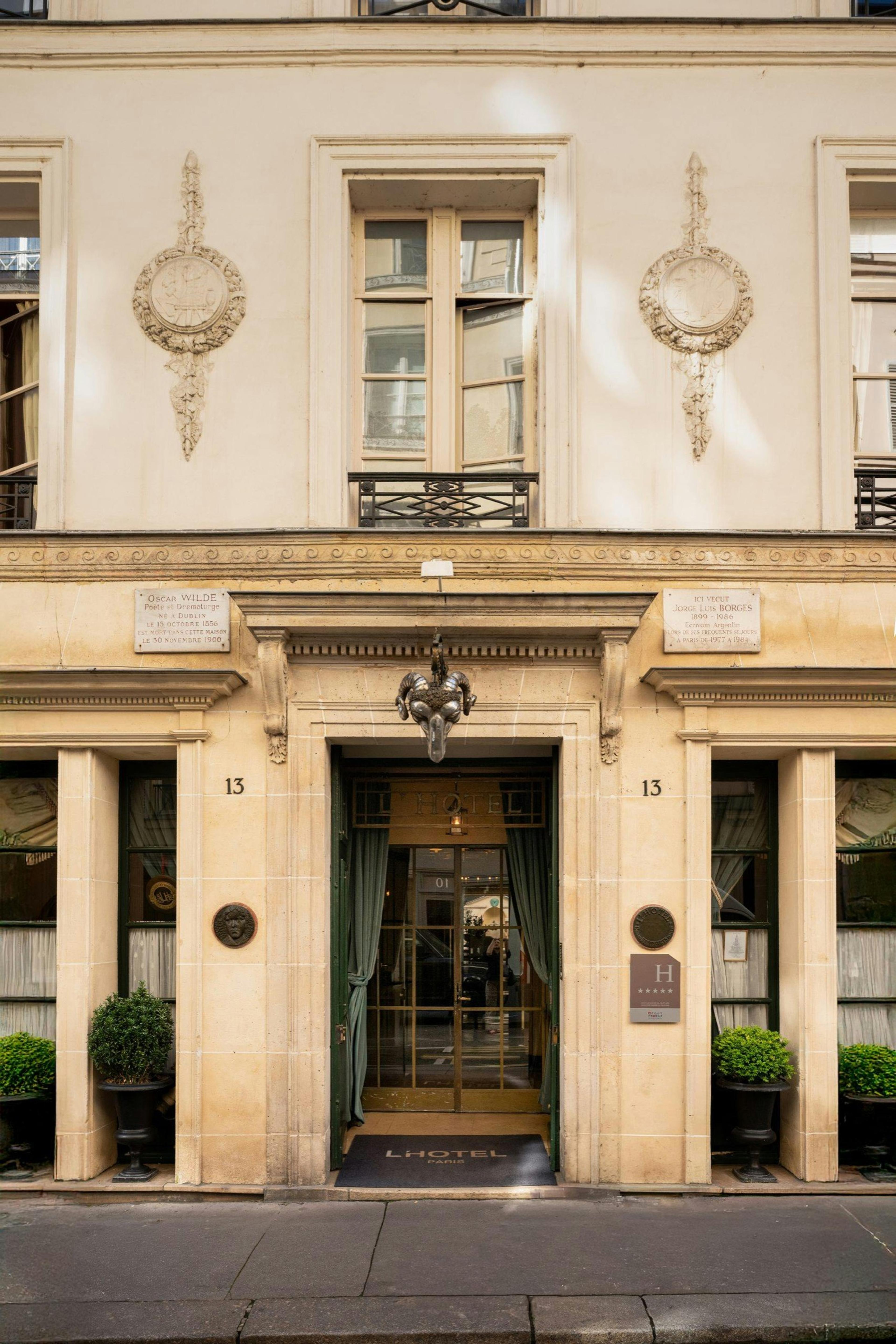 The hotel's cream stone facade and glass entrance doors at street level, flanked by potted plants and marked with the number 13 and a doormat reading 'L'Hotel Paris'.